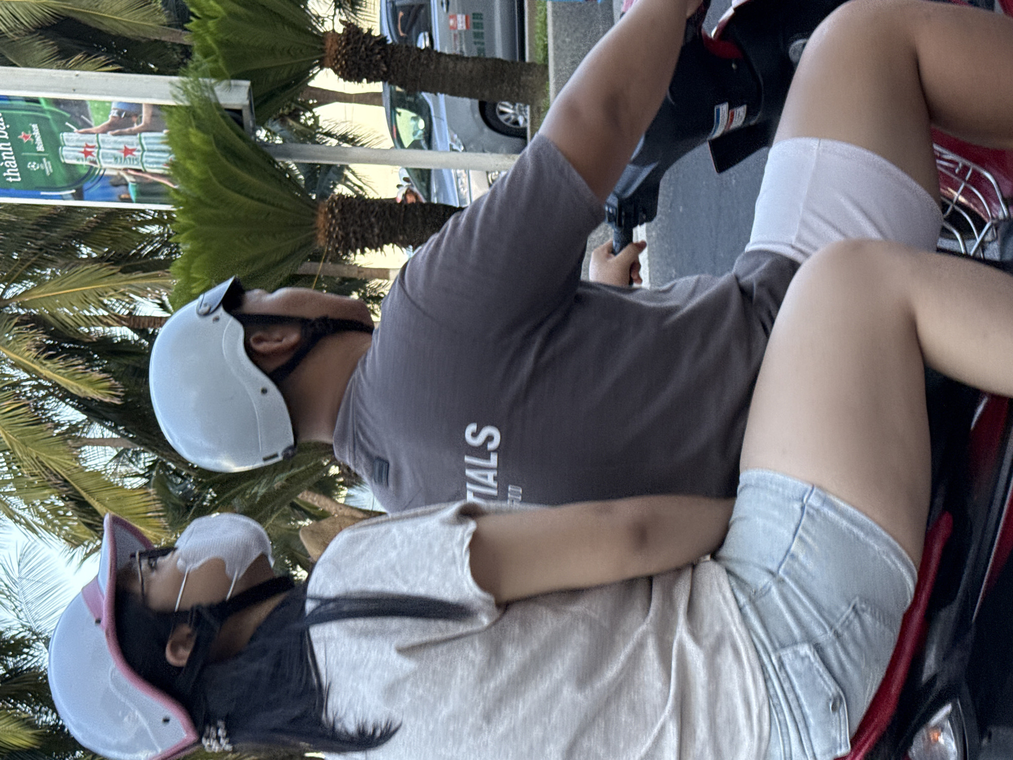 Two individuals, a woman and a man, are riding on a scooter in Nha Trang, Vietnam. The woman is in the front, wearing a light-colored t-shirt, light blue denim shorts, and a white helmet with a pink trim. She also has on glasses and a white face mask. The man is behind her, wearing a grey t-shirt with "NTIALS" printed on the back and white shorts. He is wearing a white helmet. They are riding on a road lined with palm trees and cycads. In the background, a silver car is visible, along with a billboard featuring cans of beer. The scene appears to be captured during daylight hours.