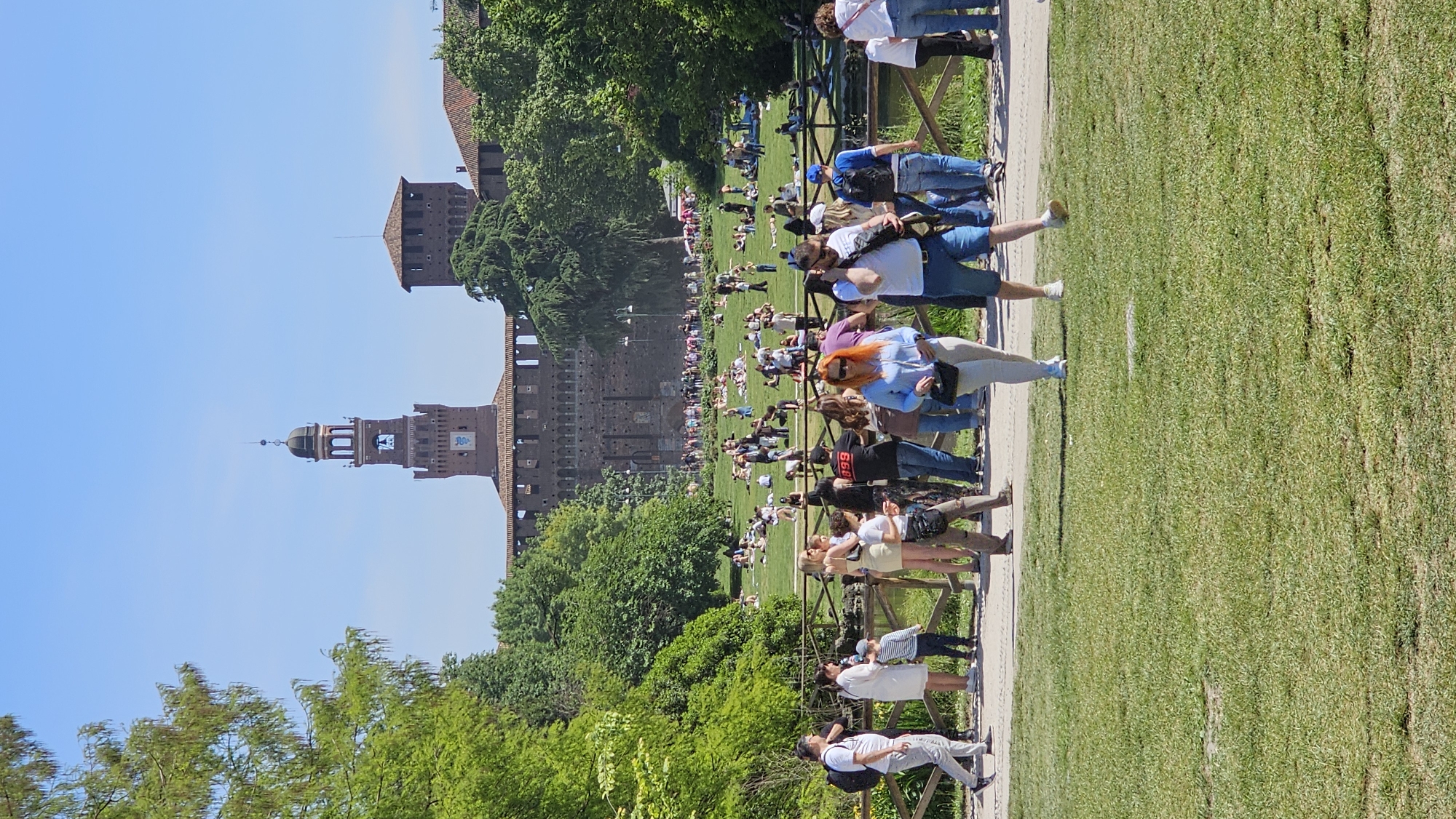 The image depicts a scene in Milan, Italy, featuring the Sforza Castle in the background. The castle is a large brick structure with towers and battlements, partially obscured by trees. In front of the castle is a large grassy area where many people are lounging and sunbathing. A path runs along the edge of the grassy area, separated by a low wooden fence. People are walking along the path, some appearing to be tourists. The sky is clear and blue, suggesting a sunny day. The foreground includes additional trees and greenery.