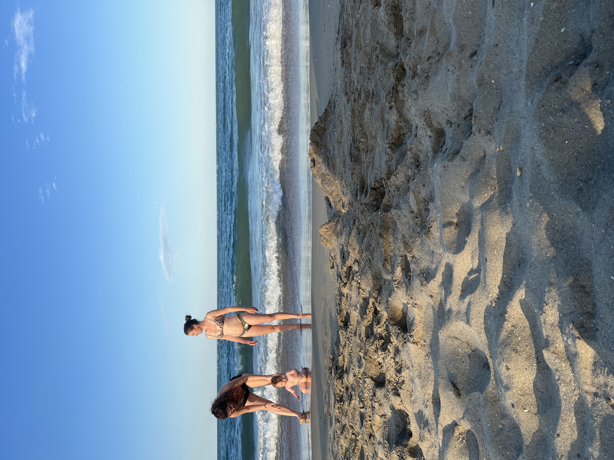 At the edge of the sea in Constanța, Romania, two women and a baby are enjoying a day at the beach. The woman on the left, wearing a dark bikini, is bending down to interact with the baby. The baby, naked and sitting in the shallow water, appears to be playing. The second woman, in a patterned bikini, stands a few feet away, looking out at the ocean. The water is calm with gentle waves, and the sky is a clear, bright blue with a few wispy clouds. The sandy beach is in the foreground, with some footprints and a small sandcastle visible.