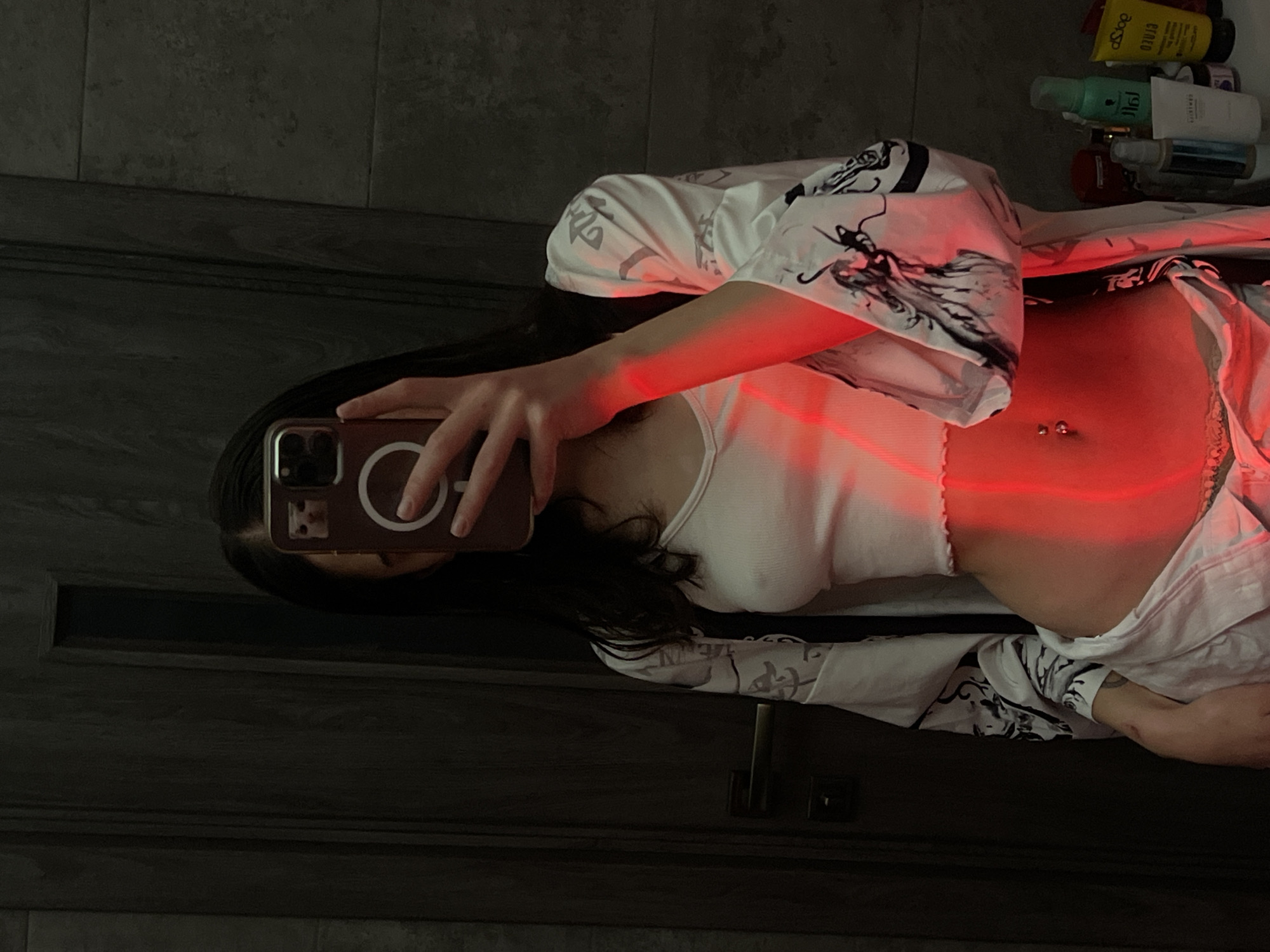 The image shows a young woman taking a selfie in a bathroom. She is wearing a white crop top and white pants, with a patterned robe or cardigan over her shoulders. She has a belly button piercing. A red light casts a glow across her midsection. On a shelf to the right, various toiletries and beauty products are visible. The background appears to be a tiled wall and a dark wooden door. The scene is consistent with a private residence bathroom. There is no specific information in the image that would allow for identification of the location within Ternopil, Ukraine.