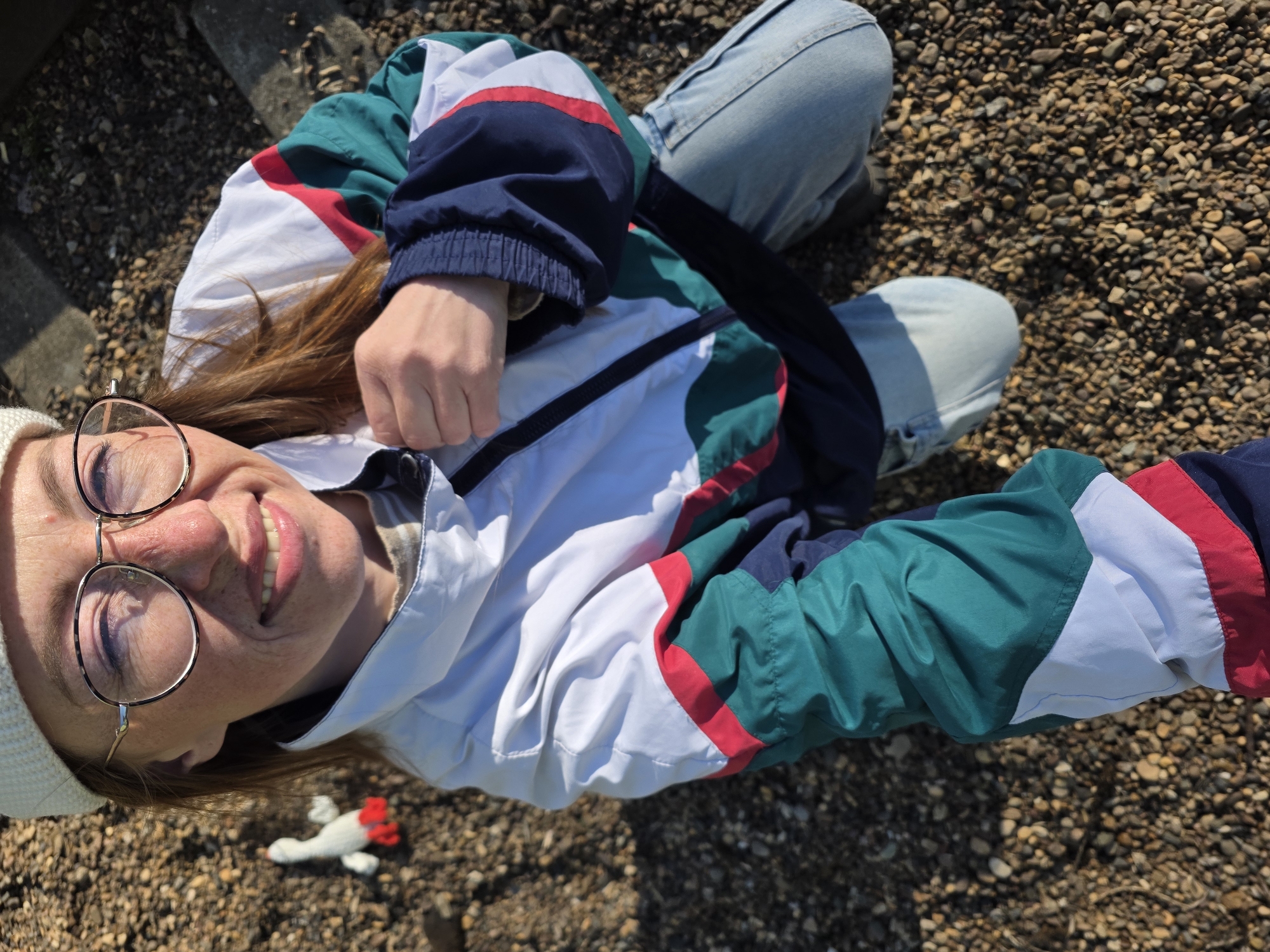 A woman with round, wire-rimmed glasses, wearing a white beanie and a 1990s-style windbreaker jacket in white, teal, navy blue, and red, sits cross-legged on a surface of small, brown pebbles. She is smiling and looking up at the camera, which is positioned above her at an angle. Part of her light-wash denim jeans are also visible. Near the woman's head, on the pebble surface, is a small, white knitted toy with red details. The location is possibly Chaikovsky, Russia based on given instruction. There are cement edging blocks partially visible in the upper left.