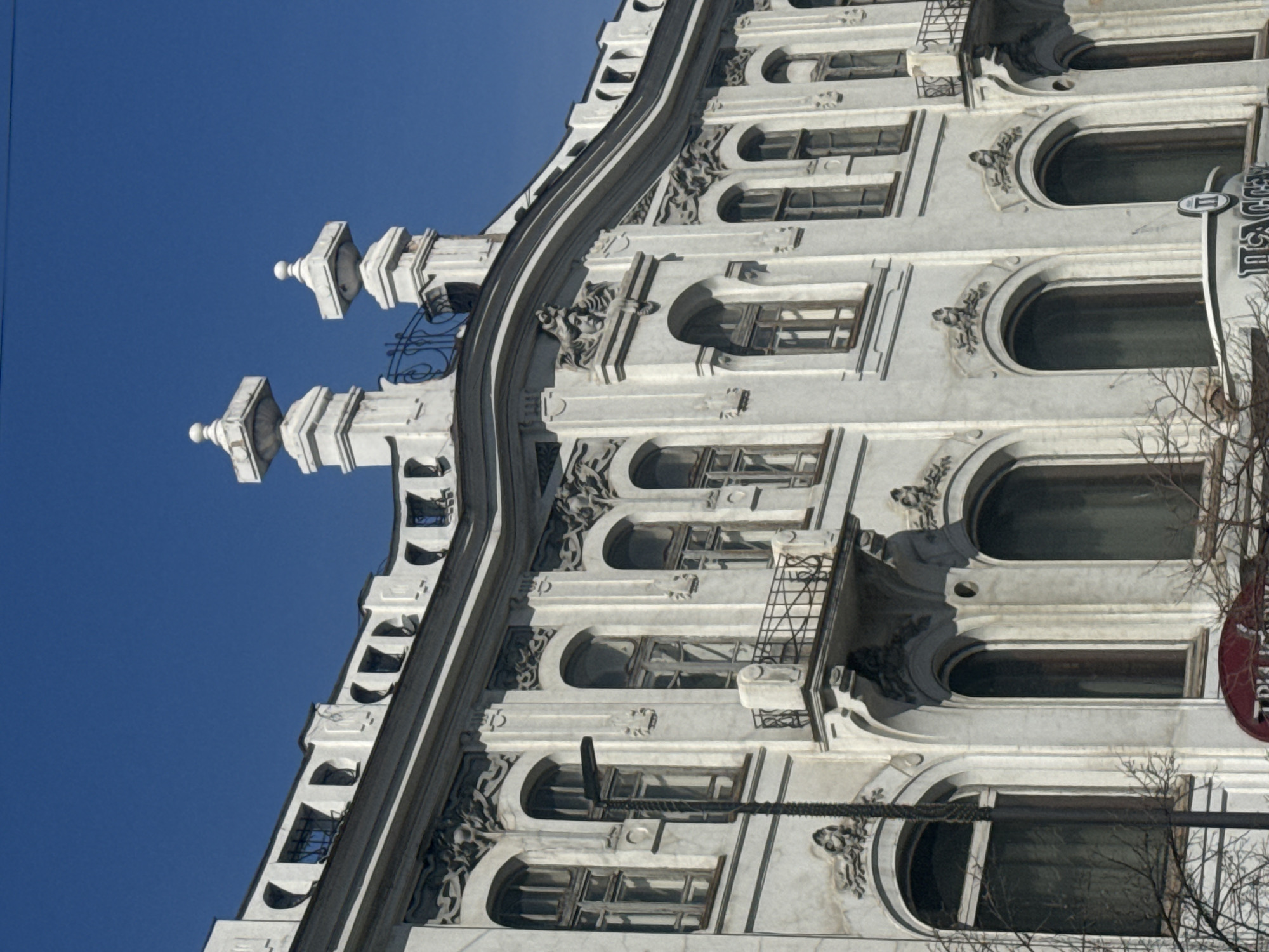 The image displays the ornate facade of a historic building in Krasnoyarsk, Russia, captured from a low, upward angle against a vivid, clear blue sky. The multi-story structure features a light-colored, possibly off-white, exterior adorned with intricate architectural details, suggesting an Art Nouveau or similar style.
The building is characterized by numerous arched windows, many of which are enhanced with decorative relief carvings above them, depicting stylized figures or floral motifs. Some upper-floor windows include small balconies with delicate wrought iron railings. A prominent, curvilinear cornice runs along the top of the facade, capped by several white, pyramidal finials with spherical tops.
The bright daylight creates subtle shadows that emphasize the building's three-dimensional elements. On the right side, bare tree branches partially frame the scene, suggesting winter or early spring. A dark cable or wire stretches horizontally across the middle-right of the frame. A vertical sign is partially visible on the building's right, displaying Cyrillic letters that appear to spell out "ПАССАЖ" (PASSAGE). No people or explicit activities are depicted, focusing solely on the architectural grandeur.