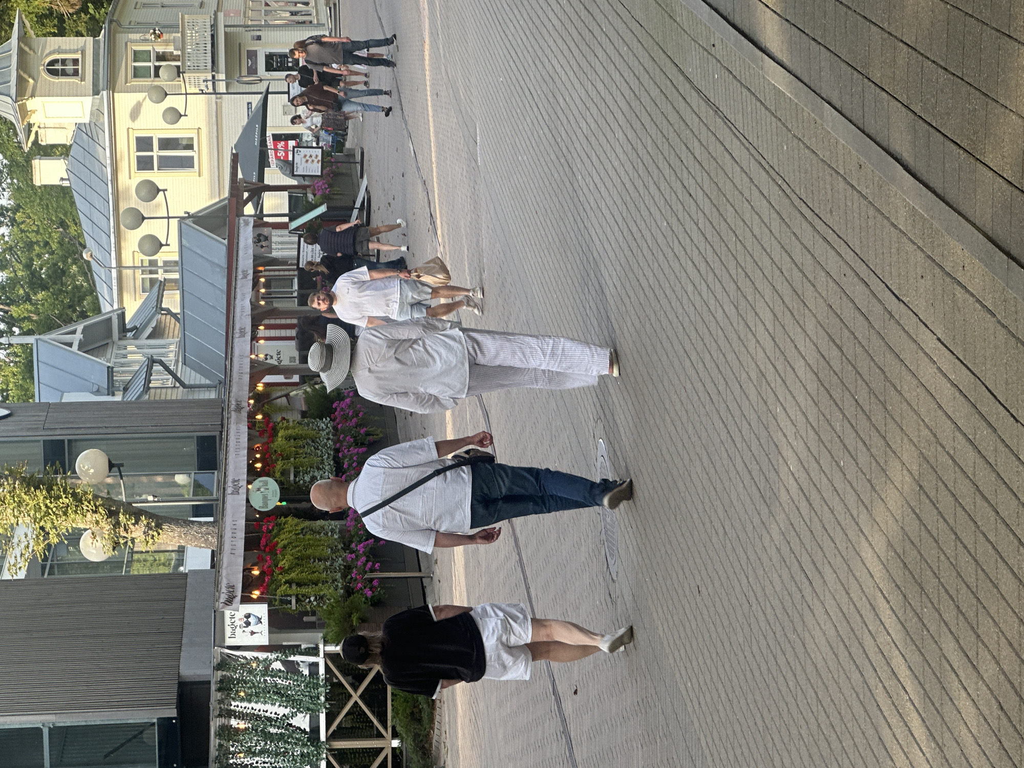 In Jūrmala, Latvia, on a paved pedestrian walkway, several individuals are seen walking away from the camera, towards a row of buildings housing shops and restaurants. The buildings feature a mix of modern and traditional architecture. On the left, a modern building with a dark, ribbed facade has large windows, and in front of it, a decorative white fence encloses plants and outdoor seating areas for a restaurant. Adjacent to this is a building with white awnings and hanging lights, displaying signage for "Baguette" and "Ristorante". Further down the street, on the right, a light yellow building with a balcony and a tower showcases a more ornate architectural style. The walkway is paved with rectangular stones laid in a diagonal pattern, with some visible grout lines and cracks. A few people are dressed in casual summer attire, including shorts, t-shirts, and light pants, with one individual wearing a wide-brimmed hat. The lighting suggests it is daytime, with shadows cast across the walkway.