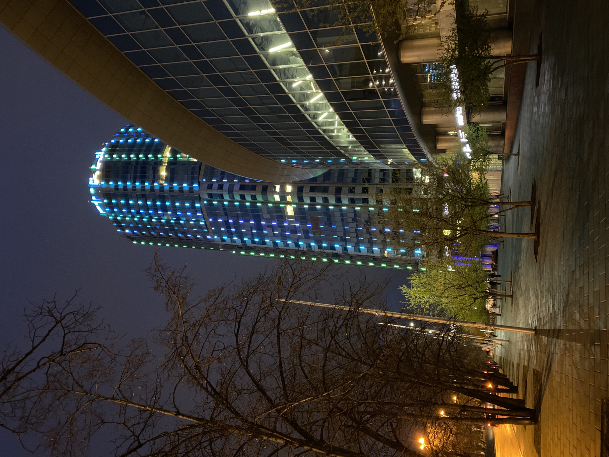 This night photograph captures a modern urban landscape in Yekaterinburg, Russia. The central subject is a tall, sleek skyscraper with a curved, glass facade, dramatically illuminated by a grid pattern of alternating blue and green lights, creating a striking visual effect. The top portion of this building features a distinct lighter-colored, curved architectural element.
Adjacent to it, another large, contemporary building with a reflective glass exterior is visible, its surface mirroring interior lights. A prominent, golden-hued architectural overhang or walkway gracefully curves across the upper part of the frame, contributing to the sophisticated aesthetic.
In the foreground and midground, a wide, paved pedestrian pathway extends into the distance, lined by a row of trees. The pathway appears damp, reflecting the warm yellow glow from several streetlights positioned along its length. Some trees in the immediate foreground are bare, their intricate, dark branches silhouetted against the dark sky, suggesting a cold season, while other trees further along the path exhibit vibrant green foliage. A few indistinct human figures can be seen walking in the distance along the illuminated path.
The sky is a deep, dark blue, confirming the nighttime setting. The scene conveys a sense of urban modernity and architectural impressiveness, bathed in the quiet ambiance of an evening. No legible text is visible within the image.