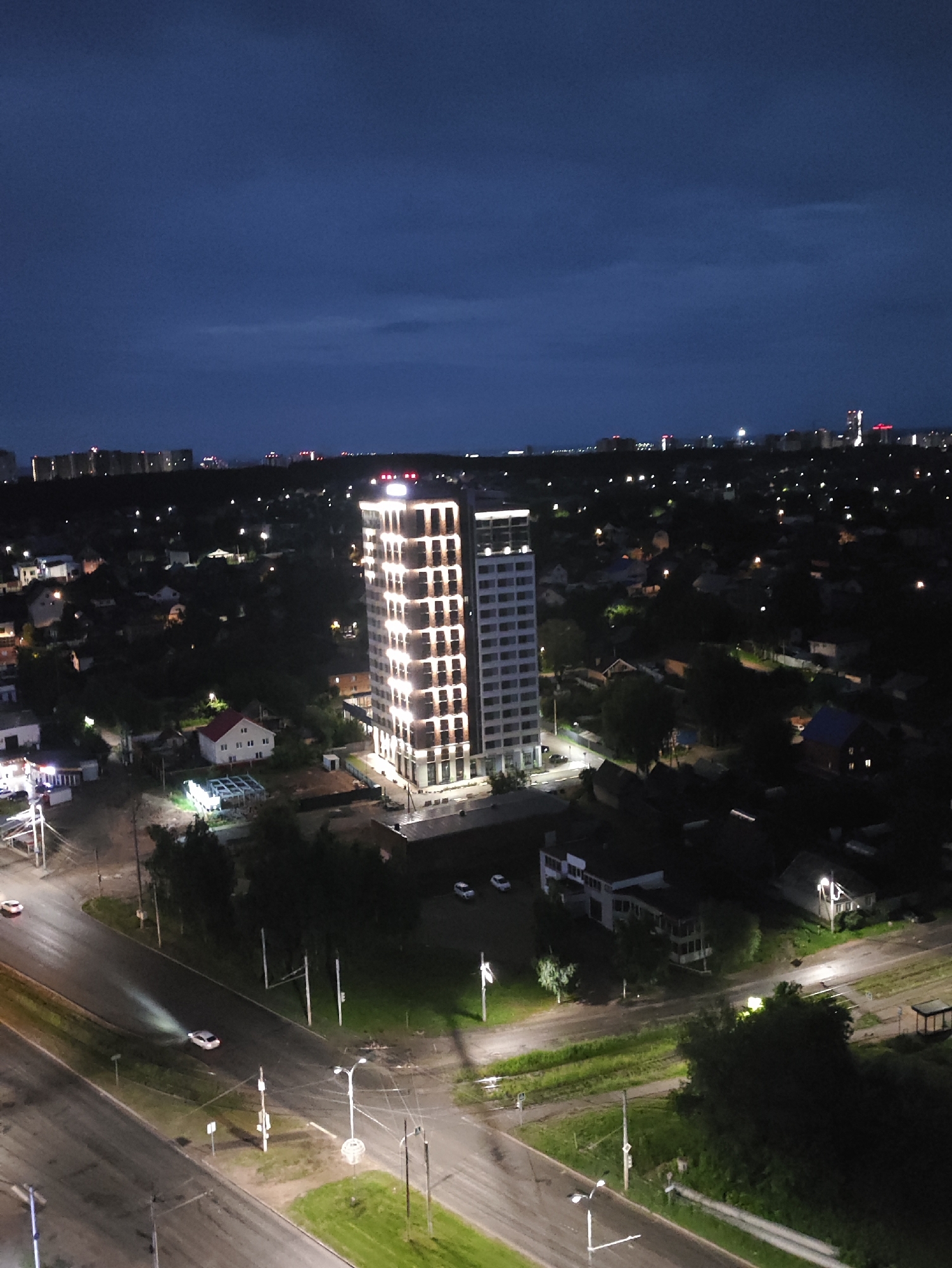 An aerial, medium-distance, night-time photograph captures the city of Izhevsk, Russia, showcasing a cityscape with a prominent modern building and a surrounding residential area. The sky is a dark, deep blue with subtle cloud formations. In the foreground, a street with two lanes is visible, with one white car in the left lane and another white car in the right lane, both appearing to be in motion. The streetlights cast pools of light on the wet asphalt surface.
Dominating the mid-ground are two tall, contemporary buildings. The building on the left is a high-rise structure with a distinctive facade illuminated by numerous vertical rows of bright white lights, highlighting its modern architectural design. Adjacent to it, to the right, is a slightly shorter, sleeker building with a lighter exterior and evenly spaced rows of windows, also illuminated from within.
Surrounding these buildings is a densely populated residential neighborhood characterized by numerous low-rise houses, many with illuminated windows. Trees and foliage are scattered throughout, contributing to the urban landscape. Further in the background, a distant skyline of more buildings and lights is visible, suggesting the broader extent of the city. The overall impression is of a well-lit urban environment at dusk or night.