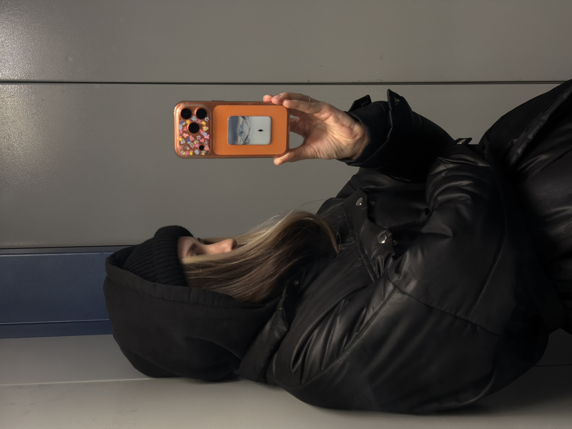 A person is lying on their side, likely on a bench, indoors, with their head towards the left side of the frame. They are wearing a black hooded sweatshirt and a black puffer jacket. The person is holding an orange smartphone case in their right hand, taking a picture or video. The background consists of a grey wall and a blue-grey panel below the wall. There is no evidence in the image to verify this photo's location.
