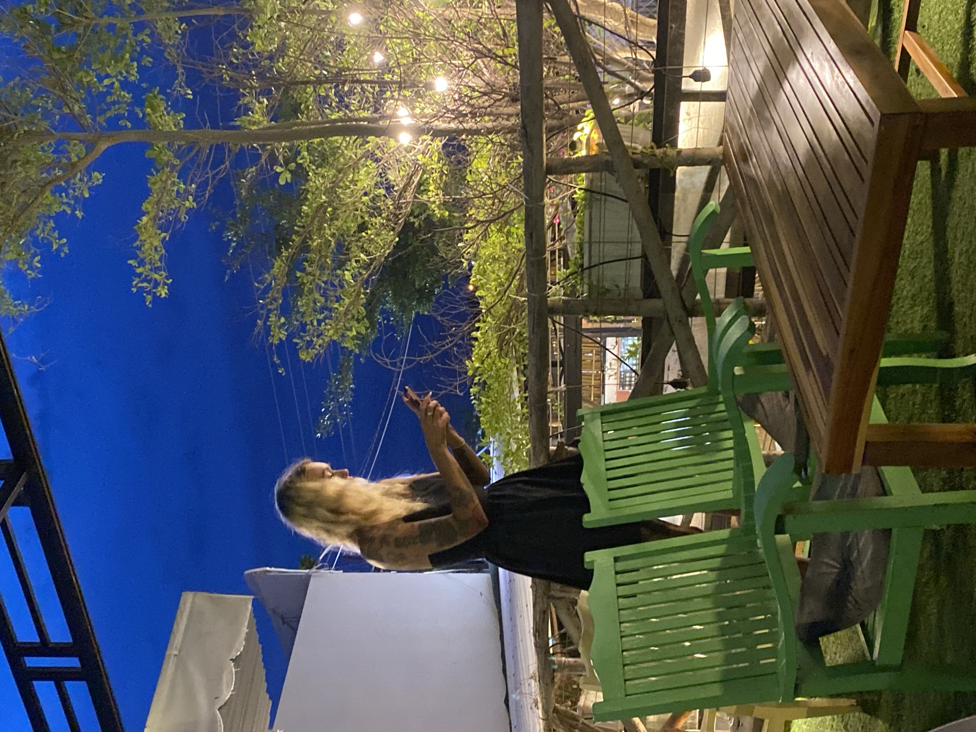A woman with blonde hair, wearing a black dress and arm tattoos, stands on an outdoor patio at night, looking at her phone. She is positioned to the left of the frame, with a white wall to her left and a wooden railing behind her. In the foreground, two bright green chairs face a wooden table, set on a grass-like surface. Behind the railing, trees with illuminated string lights are visible against a deep blue sky. The scene suggests a relaxed outdoor setting, possibly a cafe or restaurant, in Phu Thuy Ward, Vietnam.