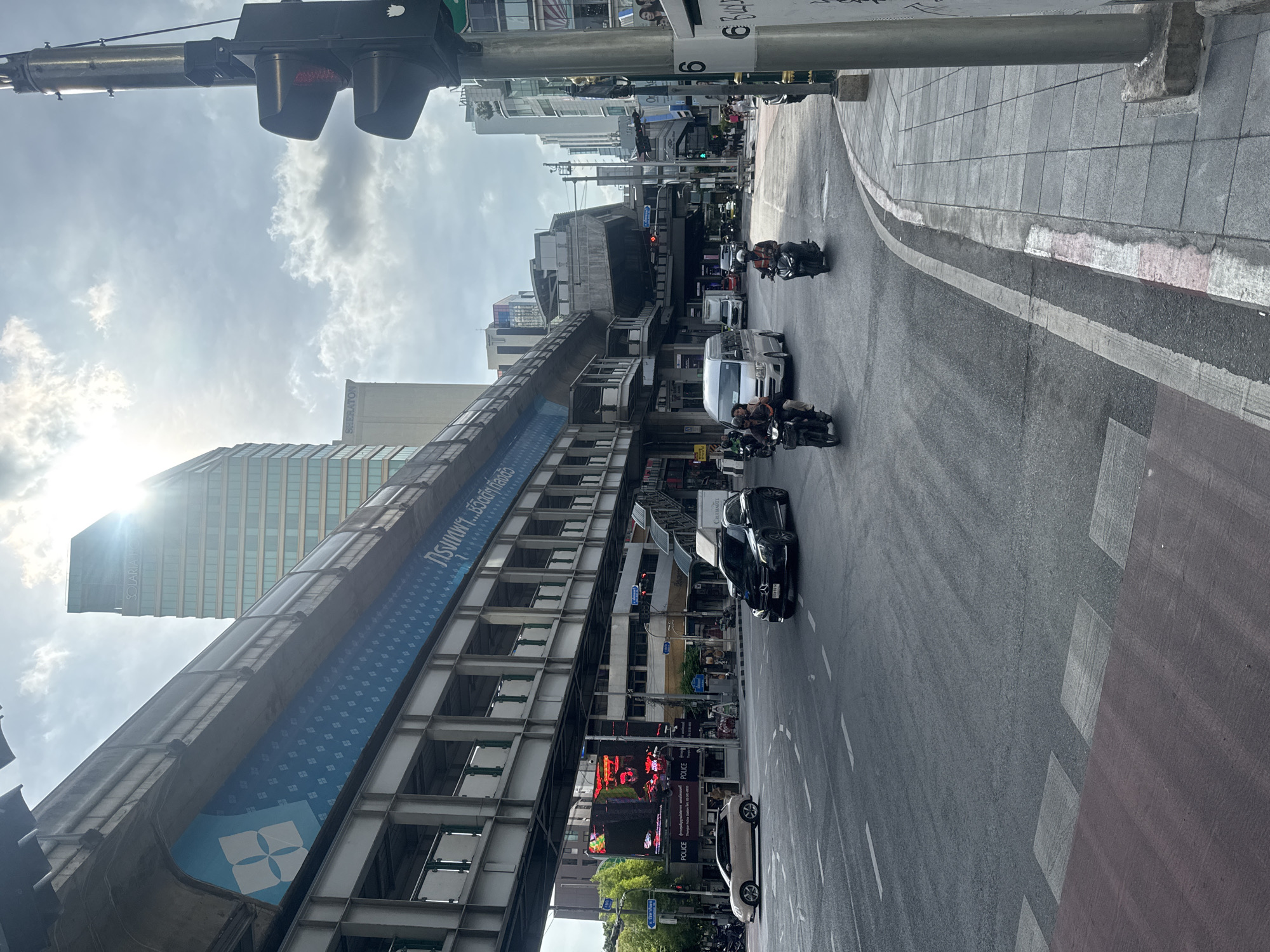 The image captures a street scene in Bangkok, Thailand, dominated by an elevated BTS Skytrain track to the left. A tall building can be seen behind the Skytrain. Below the Skytrain structure is an LED advertising screen.
The street is visible with multiple vehicles including motorcycles, a passenger van, and cars moving along the road. A traffic light is positioned in the upper left of the frame. Pedestrian crosswalks are marked on the right side of the street. The sky is overcast with bright sunlight filtering through the clouds.