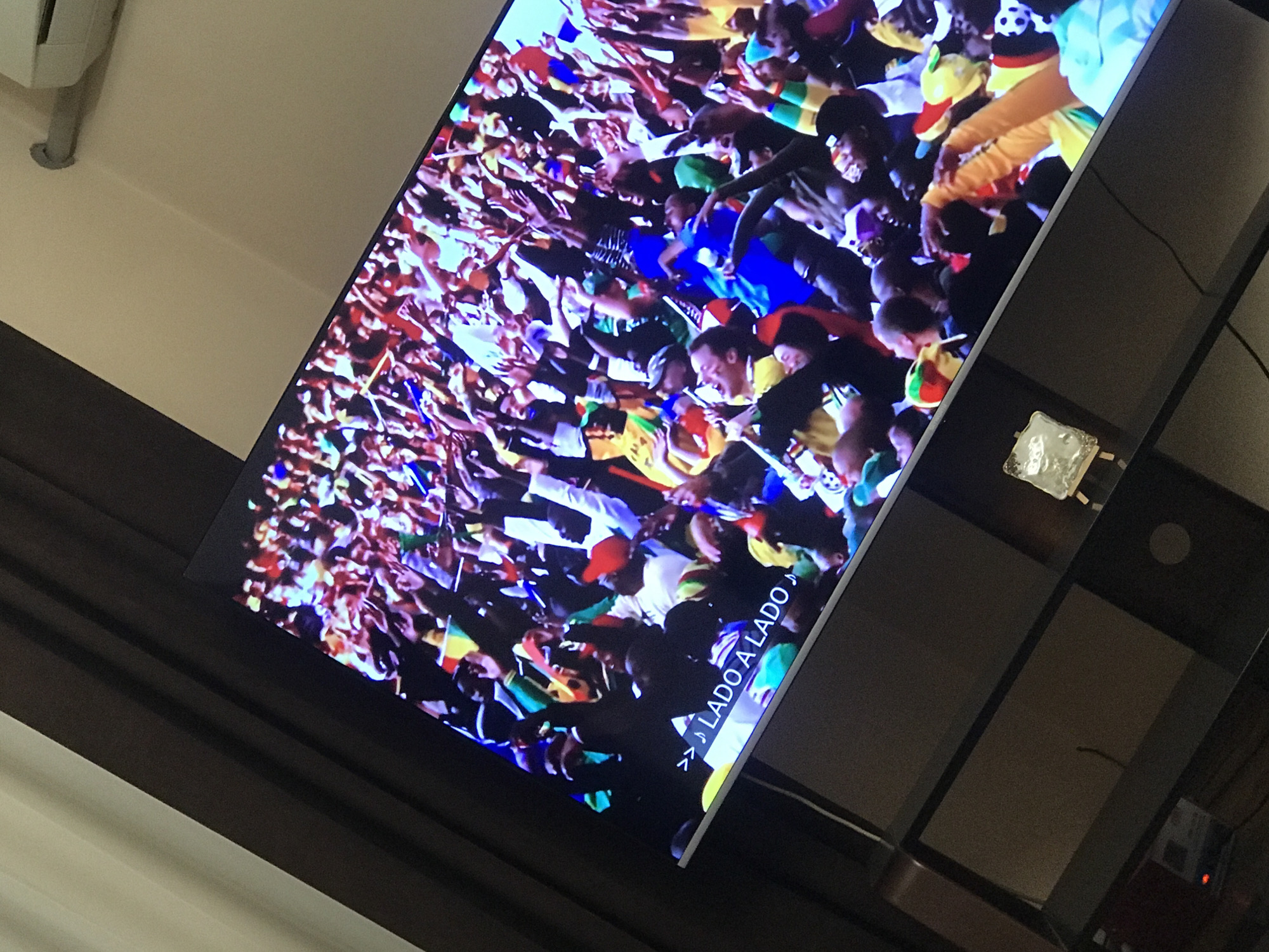 The image shows a television screen displaying a crowd of people, many of whom are wearing colors associated with the flag of Senegal (green, yellow, red). Many hands are raised in the air. The text "> LADO A LADO " is visible on the screen. The television is sitting on a black stand against a background of light colored walls and dark brown window frames. There is a lamp on the TV stand. The location is unspecified in the image and could not be determined from the available information.