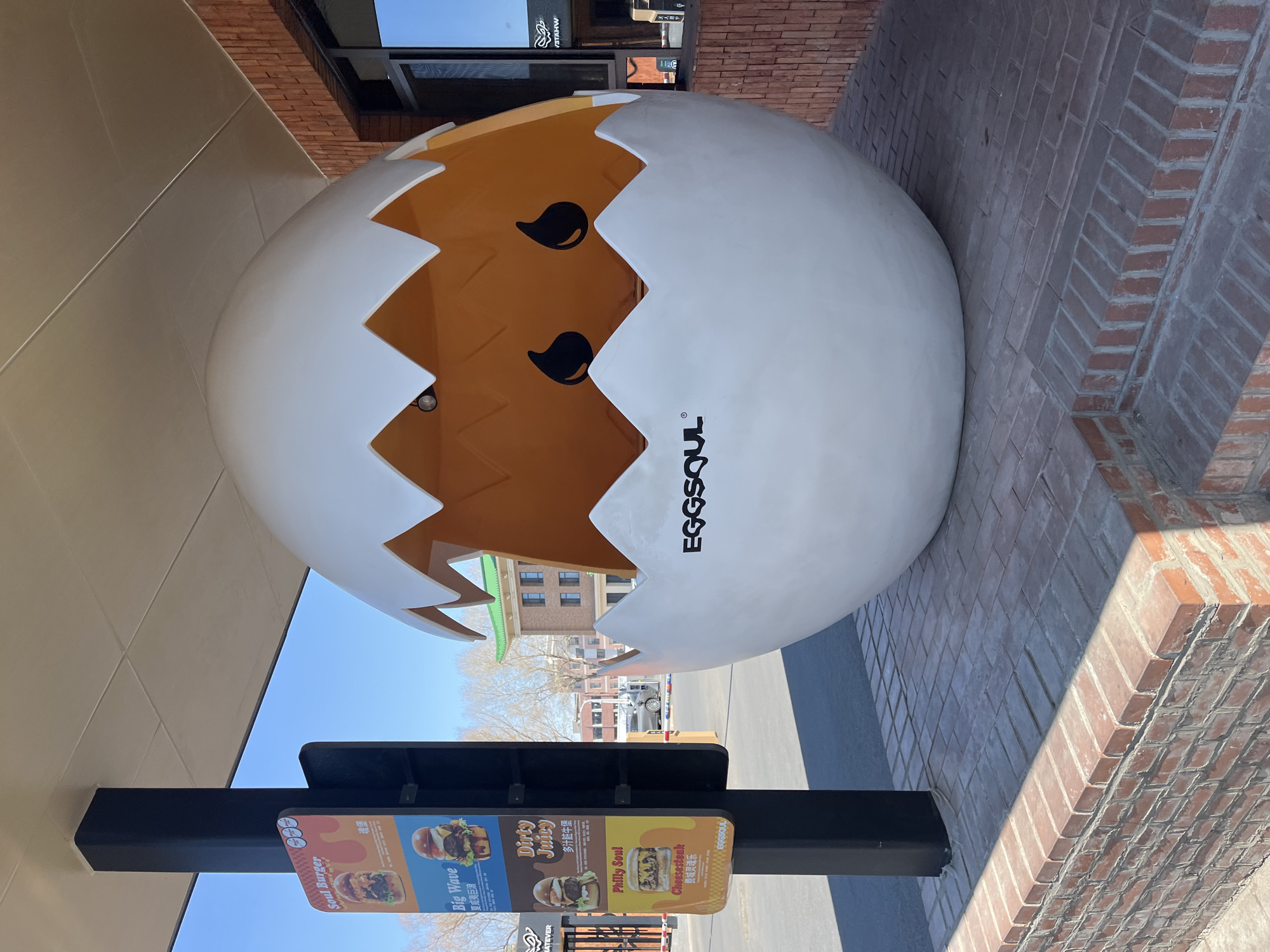 Here is a description of the image:
The image shows a large, cracked egg-shaped sculpture with cartoon eyes and the word "EGGSOUL" printed on the side. The egg is primarily white with an orange interior. A sign promoting various burgers and sandwiches, including "Soul Burger," "Big Wave," "Dirty Juicy," and "Philly Soul Cheesesteak", is located in front of the egg. The sign includes Chinese characters. The scene is outdoors, on a brick-paved area, next to a building with a beige facade. In the background, buildings are visible, suggesting an urban setting. Based on the visible signs, the location is identified as Changchun City, China.