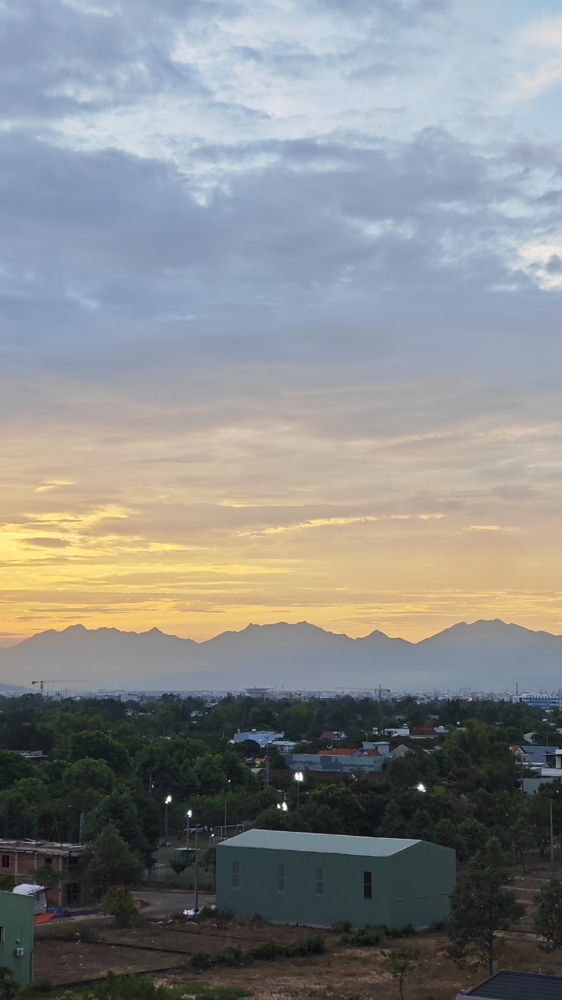 The image captures a serene dawn over Đà Nẵng, Vietnam. The sky is a canvas of soft hues, transitioning from a pale blue at the top to a vibrant golden yellow near the horizon, with streaks of grey and pink clouds painting a picturesque scene. In the middle ground, a range of silhouetted mountains stretches across the landscape, their peaks softened by the morning mist. Below the mountains, the city of Đà Nẵng unfolds with a dense canopy of lush green trees interspersed with buildings and infrastructure. Notably, a long, light green rectangular building with multiple windows stands prominently in the foreground, accompanied by construction materials and uncultivated land. The presence of streetlights, some illuminated, suggests the early morning hour. The overall atmosphere is tranquil, highlighting the juxtaposition of natural beauty and urban development.