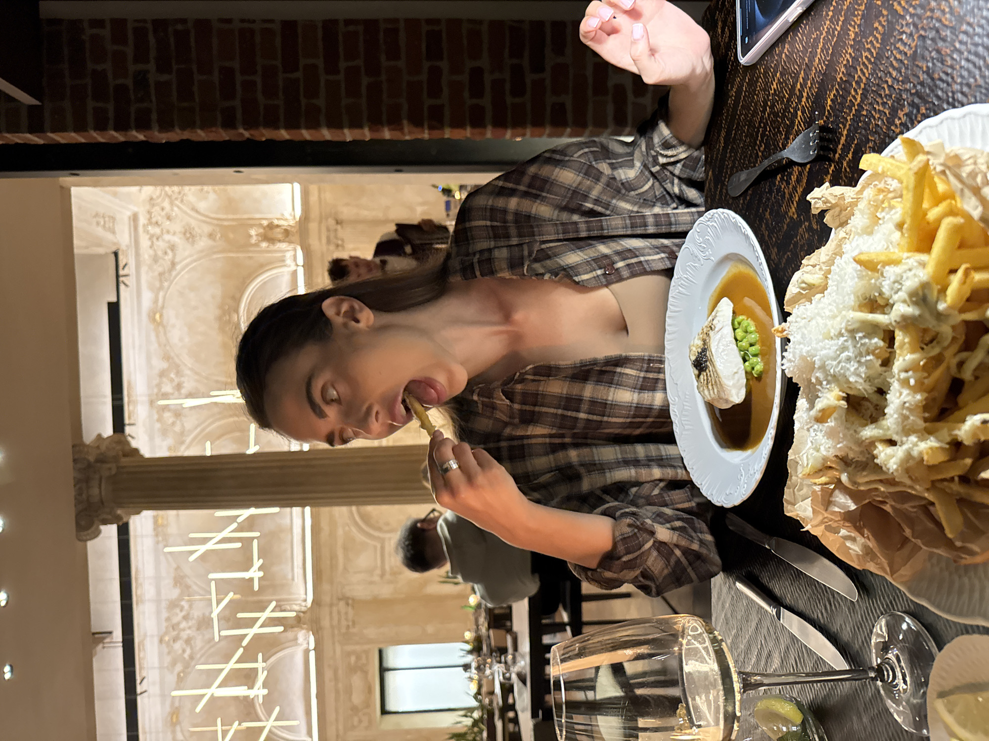 A young adult female is depicted in the foreground, in the act of putting a French fry into her widely open mouth, her tongue visible. She wears a brown and beige plaid button-down shirt over a light tan top and has a silver-colored ring on her left ring finger. Her left hand holds the fry, while her right hand, with light pink painted fingernails, rests on the table.
The dining table is dark and textured. In front of her, a white plate holds a portion of French fries served on brown parchment paper, generously topped with grated white cheese and a white sauce. To her left, another white plate contains a piece of white fish resting on green peas, surrounded by a rich brown sauce. A tall, clear stemmed wine glass containing a light liquid, two silver knives, and a small clear dish with lemon wedges are on the left side of the table. On the right, a dark fork and a smartphone in a light pink case with a dark screen are visible.
The background reveals a spacious, upscale restaurant interior characterized by classical architectural elements. High ceilings, large fluted columns with ornate capitals, and decorative arched structures with detailed molding are prominent. Modern, linear light fixtures are irregularly suspended from the ceiling, emitting a soft glow. In the mid-ground, another patron is seated with their back partially visible, and a figure resembling a server is seen standing further back. A section of exposed red brick wall is visible on the far right. The overall lighting is warm and subdued, creating an elegant ambiance.