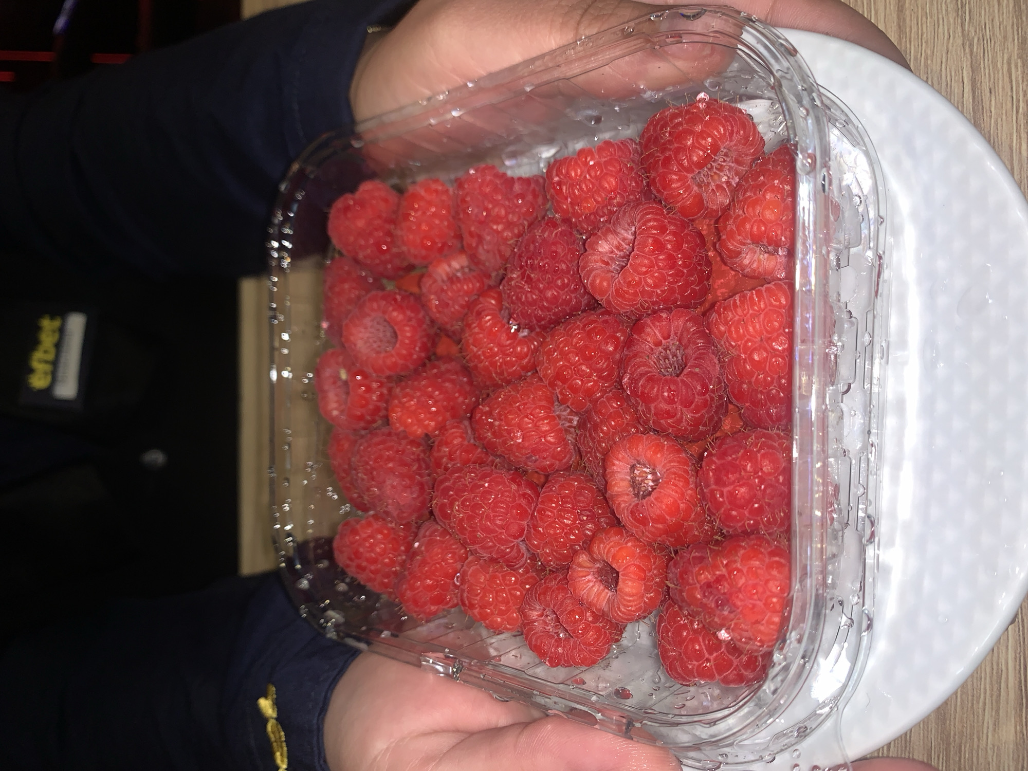 A close-up, eye-level shot shows a clear plastic container filled with fresh raspberries, held by two hands. The raspberries are plump and vibrant red, with visible water droplets on their surface and the container. The hands holding the container are lightly touching its sides, with fingers curving around the edges. The background is dark and blurred, with a hint of a person wearing a dark jacket with a yellow logo on the upper left side. The setting appears to be indoors, possibly a home or a business. Given the presence of a "efbet" logo, a prominent betting company in Bulgaria, and the visual style, the image is likely from Burgas, Bulgaria, a city where efbet has a strong presence and is a common sponsor for local establishments and events. The overall scene suggests a casual moment of presenting or preparing the fruit.