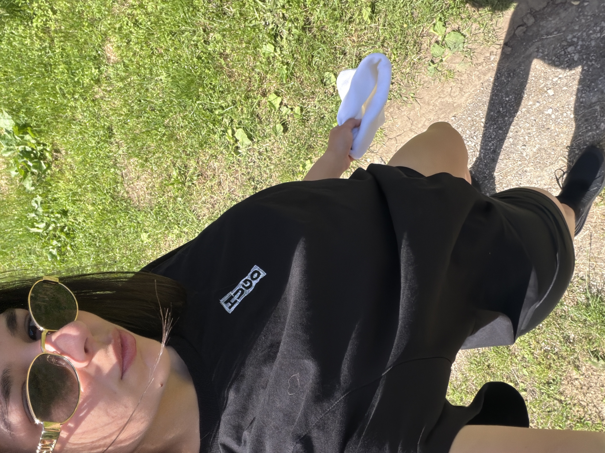 A person is standing on a path next to a grassy area. The person is wearing sunglasses and a black t-shirt with a "HUGO" logo. They are holding a white object in their right hand. Their right leg and foot are visible, and they are wearing a black shoe. Their shadow is visible on the path. The location is likely an outdoor space with grass and a path in a sunny environment. There is no information in the image to indicate the location is in Armenite, Bulgaria.