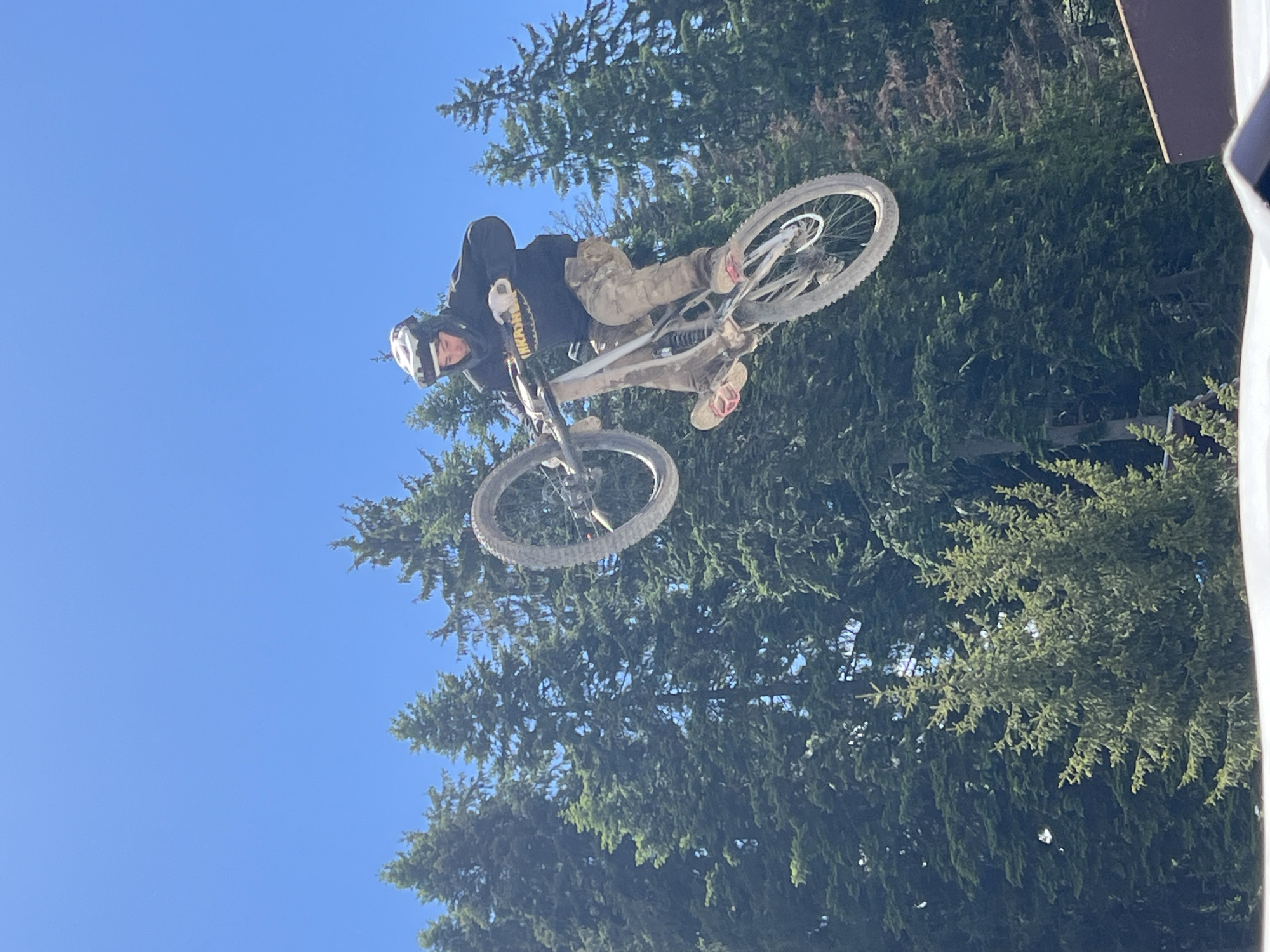 A male mountain biker, wearing a helmet and a black hoodie with yellow lettering, is captured mid-air performing a jump against a clear blue sky. He is on a dirt bike with a silver frame, and he is wearing camouflage pants and red-soled sandals. The background consists of a dense forest of tall evergreen trees. The image appears to have been taken outdoors on a sunny day. The location is not Samokov, Bulgaria.