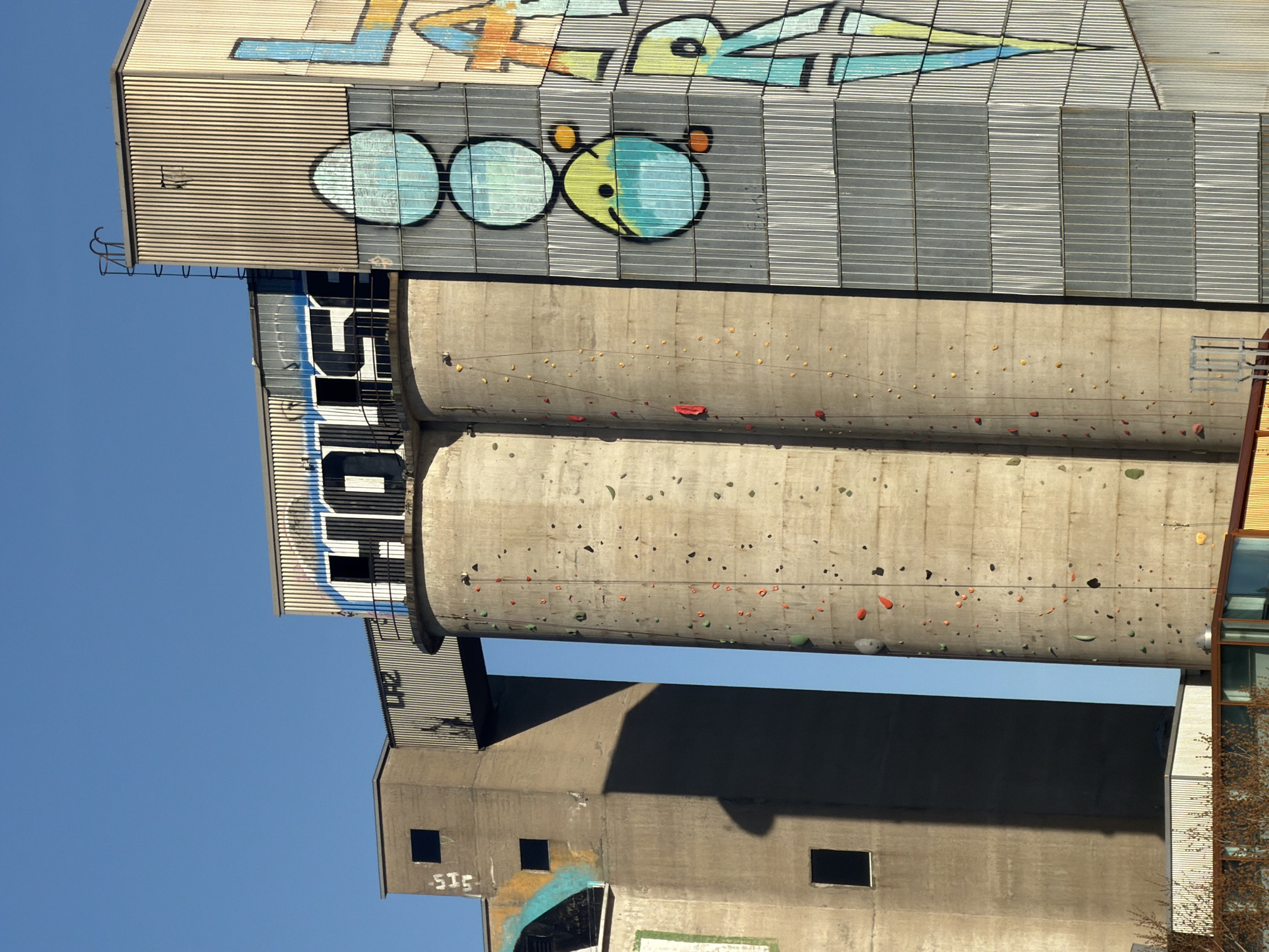 A low-angle shot shows a large concrete industrial structure against a clear blue sky. Graffiti adorns the upper parts of the structure, including a stylized depiction of a face or a series of circles resembling eyes. A section of the concrete structure is equipped with climbing holds, indicating its use as a climbing wall. There are no visible people or actions in the frame. The industrial building is located in Montreal, Canada.
