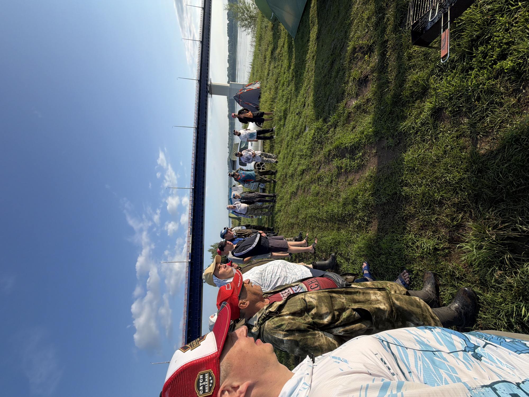 On a grassy bank near the river, under a bright blue sky with scattered clouds and a large bridge in the background, a group of men stand in a line, facing right. The men are dressed casually in t-shirts, hats, and camouflage clothing, with some wearing boots and others sandals. A few cars and tents are visible behind the group, suggesting a campsite. A barbecue grill is set up in the foreground. The scene appears to be an outdoor gathering or event. The location is identified as Tarasovo, Russia.