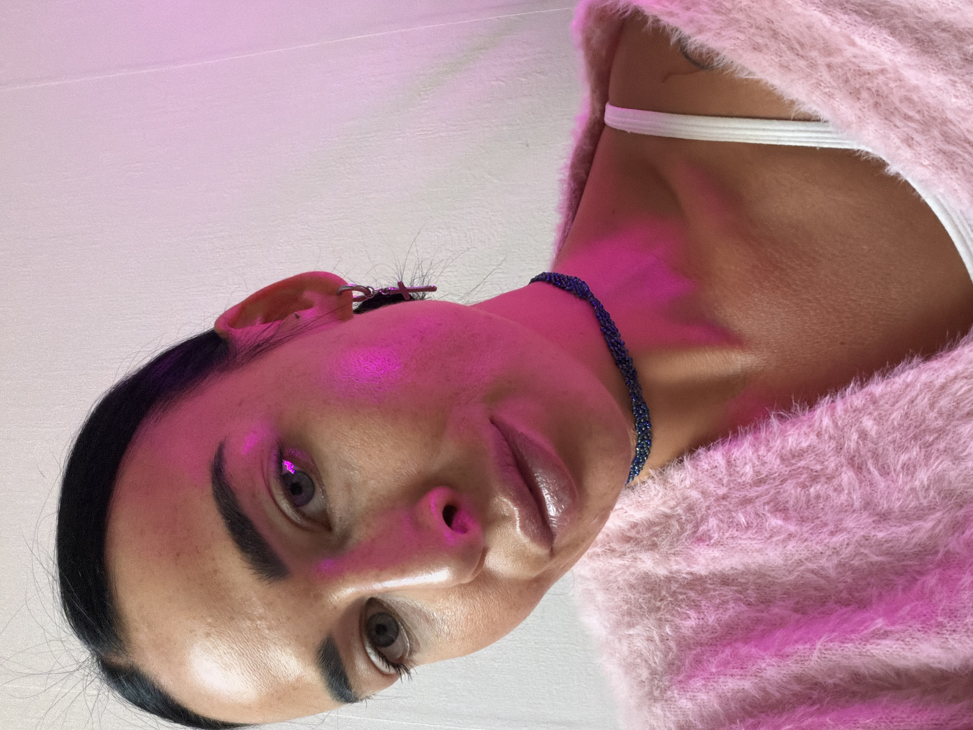 A person is positioned in a close-up, upward-facing view. The person has dark hair, defined eyebrows, light-colored eyes, and appears to be wearing a fuzzy, light pink sweater, along with a white undergarment strap visible near the neck. A dark blue beaded necklace adorns the neck. The face is illuminated by a pink light source. The background is a pale or white surface. No specific location in Saint Petersburg, Russia, can be determined from the provided image.