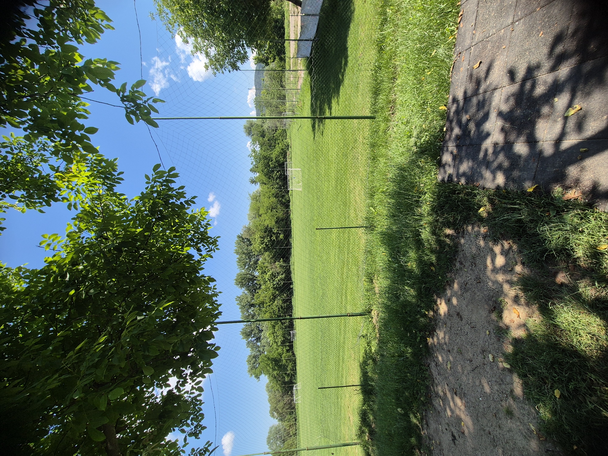 This is a medium-shot, eye-level, outdoor photograph taken on a sunny day in the town of Debelets, Bulgaria. The foreground shows a patch of sandy ground with green grass and dappled sunlight from overhanging tree leaves. A dark gray paved walkway runs along the bottom right edge of the frame. A green net fence, supported by thin green poles, separates the foreground from a large, vibrant green soccer field. In the distance, on the soccer field, are two white soccer goals. The field is surrounded by a line of lush green trees. The sky is a clear, bright blue with a few scattered white clouds. The overall impression is of a sports field on a clear, pleasant day.