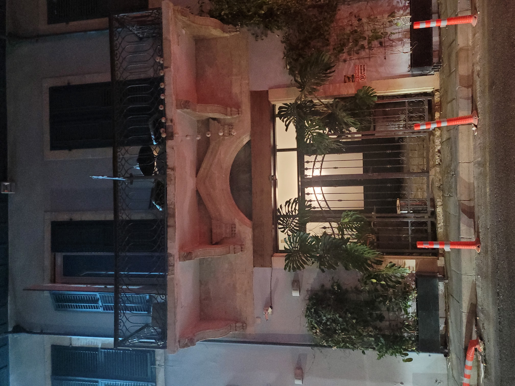The image shows the facade of a building at night in Limassol, Cyprus. The building has a balcony with ornate railings on the upper floor, supported by decorative stone brackets. Below the balcony, there is a large arched entryway with a double door made of wrought iron, flanked by potted plants, including a large Monstera. The entrance is illuminated from within. Orange traffic cones are visible on the sidewalk and street in the foreground. A sign with Greek text and a red circle with a diagonal line through it is attached to the wall to the right of the entrance. The overall scene suggests an urban environment at night, possibly a restaurant or commercial establishment given the decorative entrance and plants.