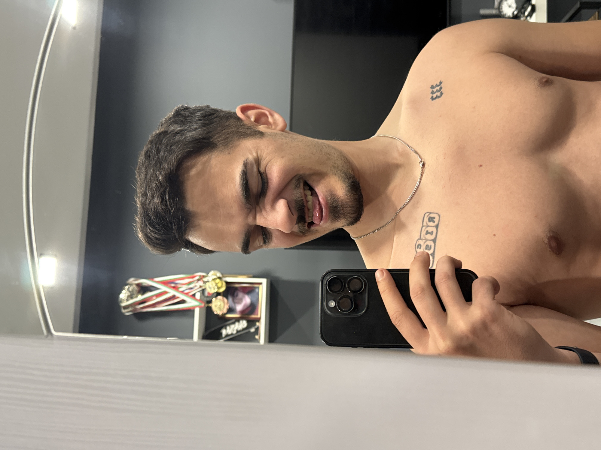A selfie captured in a dimly lit room, likely in Sofia, Bulgaria, given the medals and framed photo in the background, which may be related to local sports or events. The man is positioned horizontally, partially shirtless, showing tattoos on his chest and collarbone. He is holding a black smartphone, taking the selfie, with his face tilted and a slight smile. He wears a thin silver chain around his neck. Behind him, a mirror reflects a blurred image of the room and some decorative items. The foreground features a white surface, likely a table or shelf, on which the phone rests.