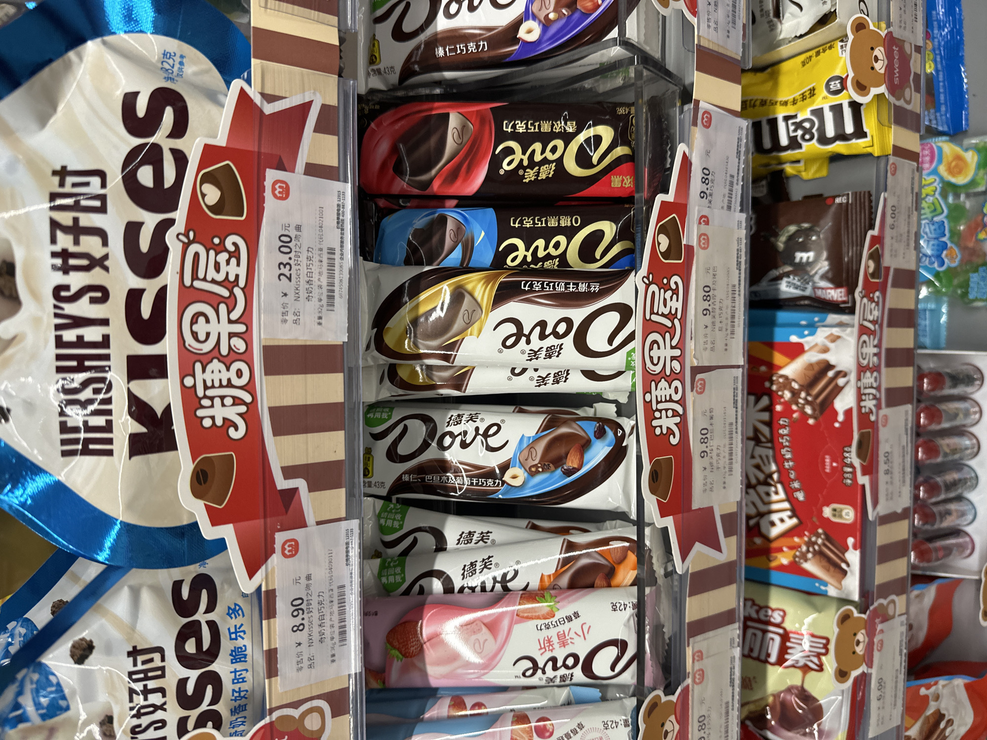 The image shows a shelf of chocolate products in a store in what is assumed to be Chongqing High-tech Zone, China, based on the Chinese characters and product labeling. The primary brands featured are Hershey's Kisses and Dove chocolate bars. Hershey's Kisses bags are prominently displayed on the left. A variety of Dove chocolate bars are neatly arranged in a clear plastic display case. The labels on the Hershey's and Dove products display prices and product information in Chinese. Adjacent to the Dove bars are products like M&M's. Each product is clearly labeled with price tags, including the brand name, product name, weight, and price in Yuan.