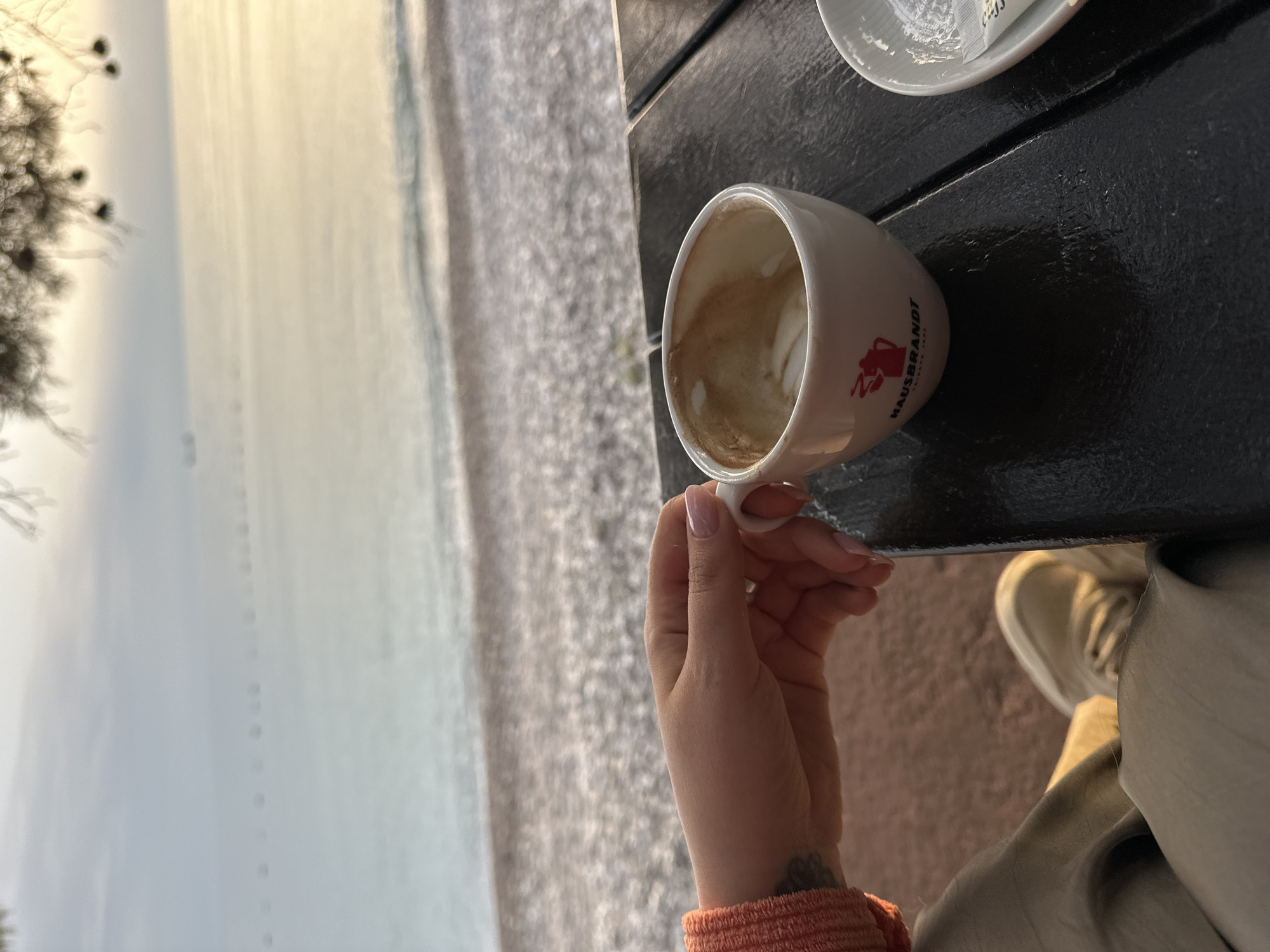A woman's hand is holding a coffee cup on a black table next to a beach. The coffee cup is white and has a logo on it. The woman is wearing a peach sweater. The beach is rocky and the water is calm. The scene is in Bar, Montenegro.