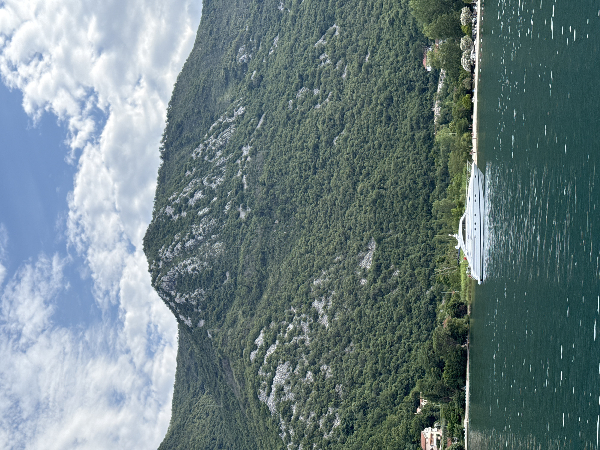 A white yacht is anchored in the foreground of a scenic body of water, its bow facing left. The water is a deep green with ripples and small white disturbances. In the background, a verdant, tree-covered mountainside rises dramatically, dotted with patches of gray rock. Along the base of the mountain, a narrow strip of sandy beach is visible, lined with white umbrellas and people. Several buildings, including houses with red roofs, are nestled amongst the trees on the slopes above the beach. The sky above is a bright blue, filled with fluffy white clouds. The scene is consistent with a view of the Bay of Kotor near Morinj, Montenegro, a popular tourist destination known for its natural beauty and coastal villages.