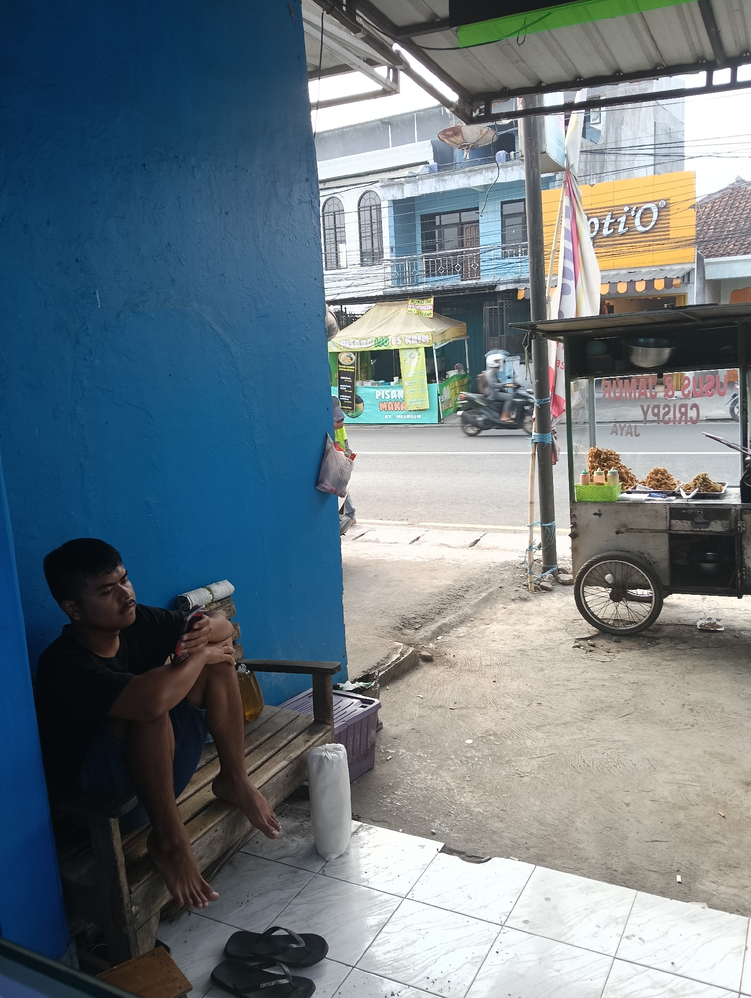 A young man with dark hair and a mustache sits on a wooden bench against a bright blue wall, looking down at his phone. He wears a black t-shirt and blue jeans, with his legs crossed and bare feet resting on the ground. To his right on the bench, a yellow bottle is visible. In front of the bench, on a tiled floor, are two pairs of black flip-flops. To the right of the bench, a white cylindrical object is propped against the wall. The scene appears to be outdoors, possibly in front of a shop or food stall. In the background, a street is visible with a food cart on wheels selling fried food, a motorcycle driving past, and buildings with yellow and blue facades. A white banner hangs vertically from a pole in front of the buildings, and a yellow sign with the text "Opti'O" is also visible. The environment suggests a commercial area in Desa Cipakat, Indonesia, likely during the daytime.