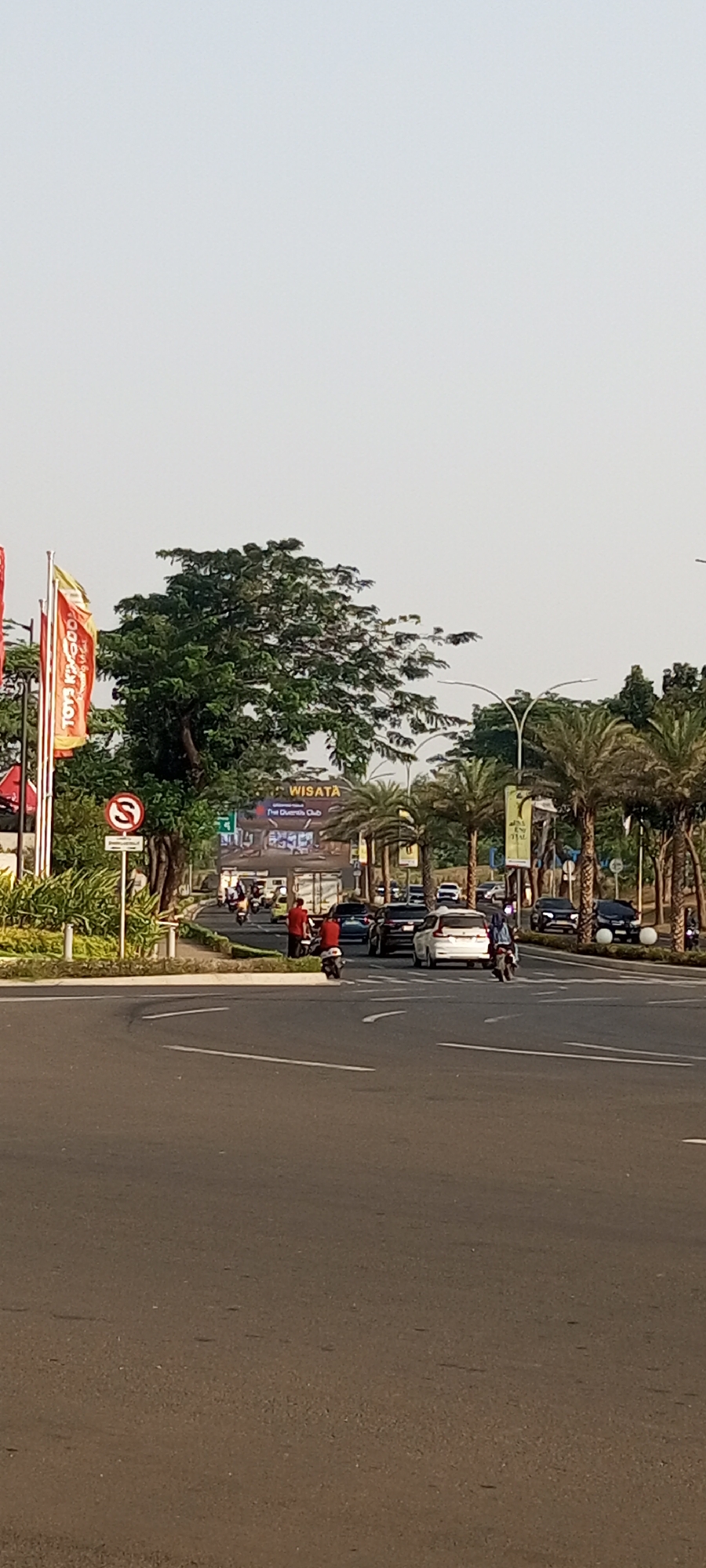 The image shows a street scene in Lambangjaya, Indonesia, with a clear sky. In the foreground, an asphalt road curves to the left. A white road marking indicates the lane division. On the right side of the road, there are several cars, including a white SUV in the middle. Palm trees line the side of the road. A billboard with the word "WISATA" and other text is visible in the background. On the left side of the road, there are flags and a signpost with a red circular sign containing a white "S." Two men in red shirts are standing near a motorcycle, and another person on a motorcycle is visible further down the road. There are also several other vehicles, including cars and motorcycles, on the road. The overall scene depicts a typical urban street with moderate traffic.