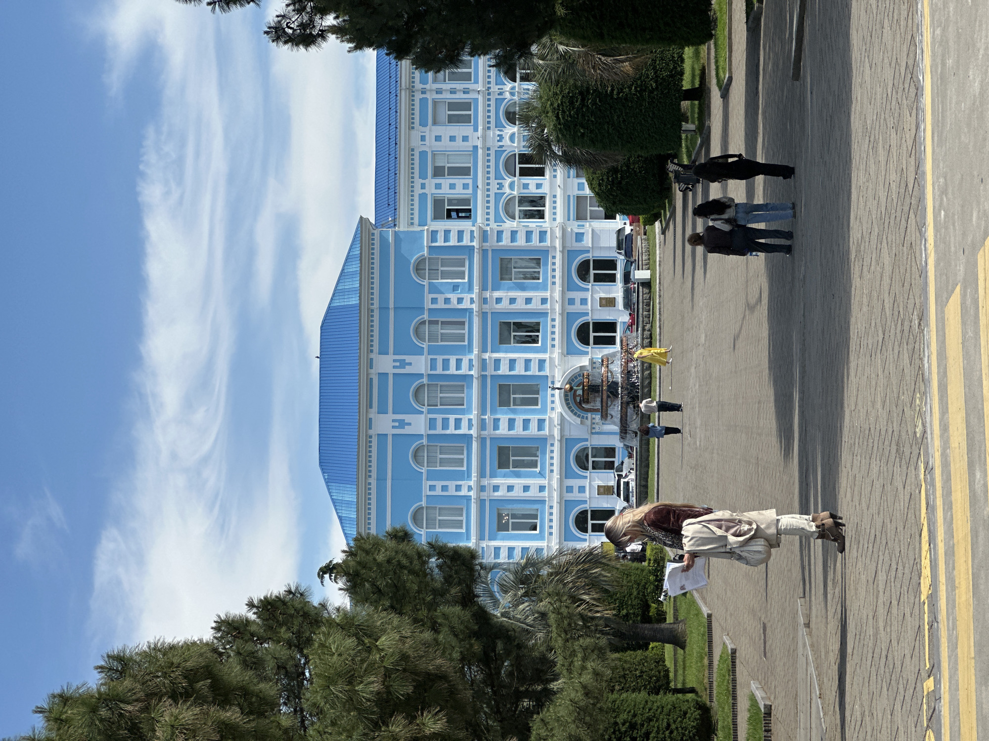 The image shows a scene in Batumi, Georgia, with a blue and white building visible in the background. The building has a distinctive architectural style. In the foreground, there is a woman standing on a paved area, holding papers. There are trees and greenery on the left side of the image, and other people walking in the background. The sky is blue with some white clouds. The image appears to be taken on a sunny day.