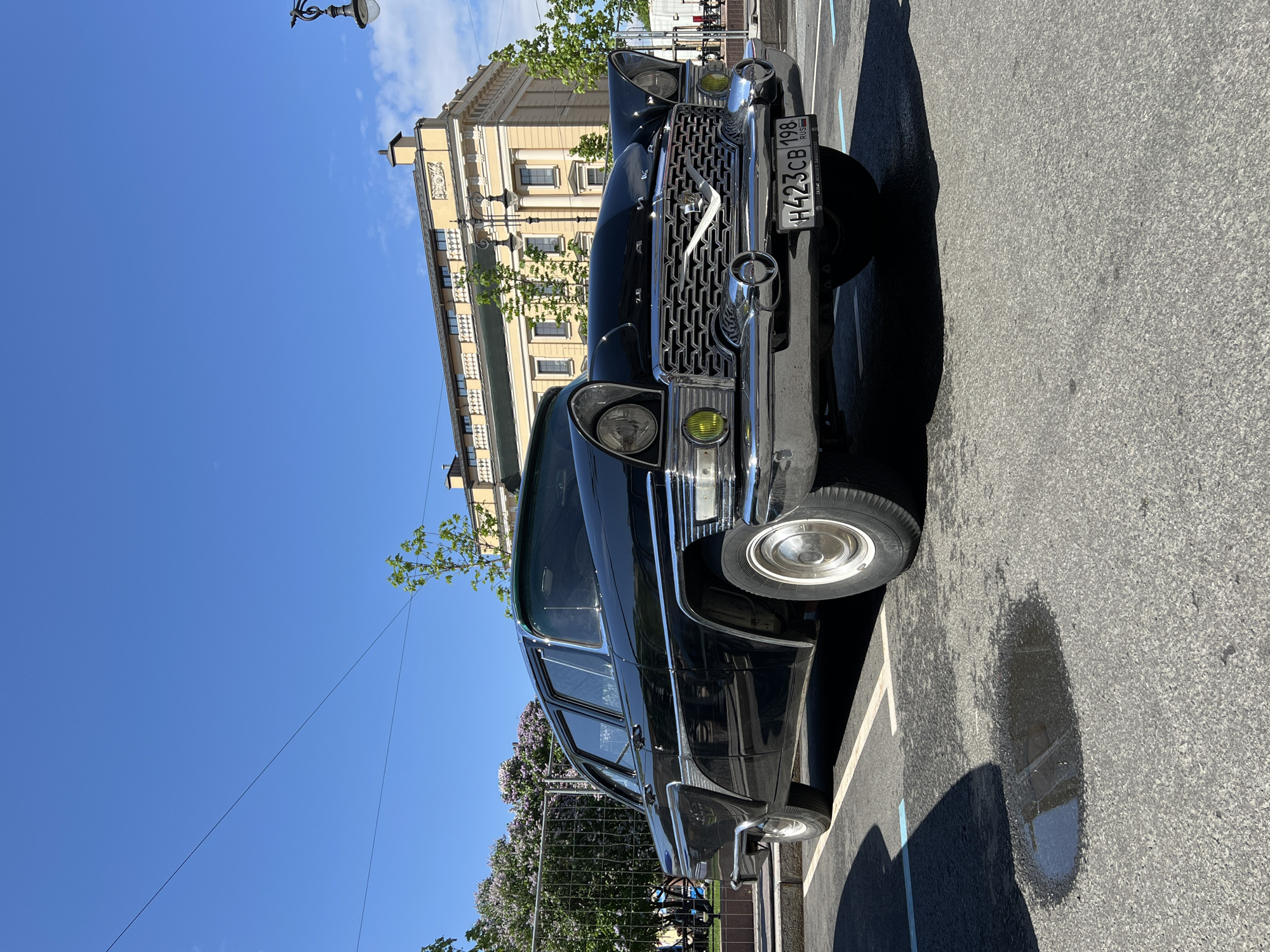 A black GAZ-21 Volga car is parked on a paved road in Saint Petersburg, Russia. The car is facing forward, with the front grill and headlights visible. The license plate reads "H423CB 198 RUS." Behind the car is a beige building with symmetrical windows and a visible lamppost. The sky is clear and blue. A small patch of water is visible on the pavement near the car. Lilac bushes are visible in the background, behind a metal fence.