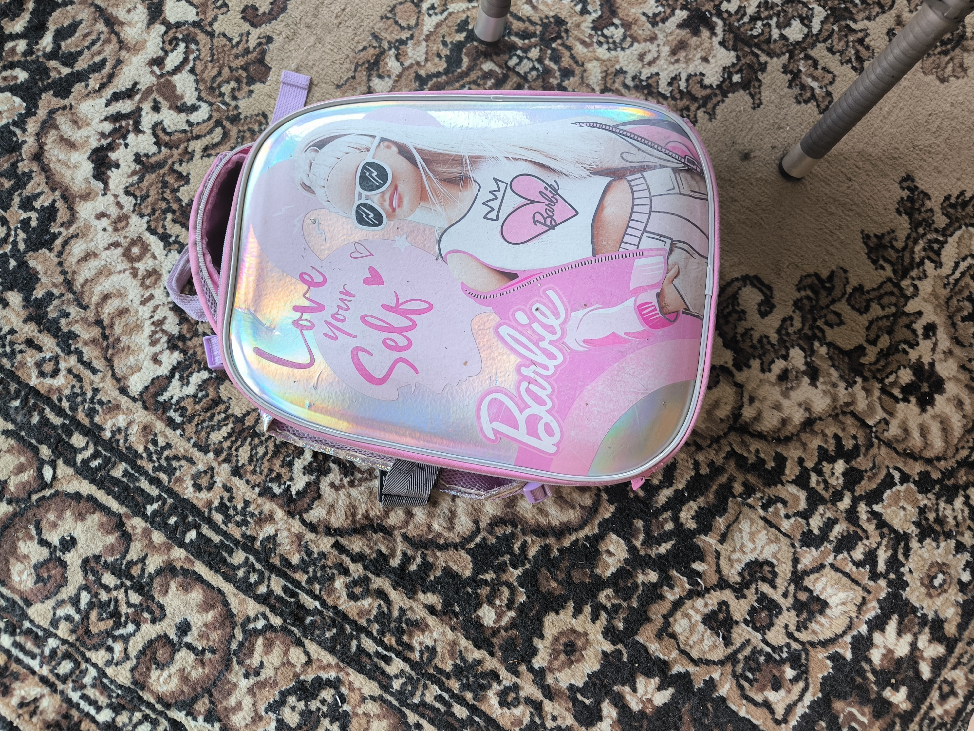 A pink, iridescent backpack with a Barbie graphic and the phrase "Love Your Self" is lying on a patterned rug. The rug has a beige background with dark brown and black intricate designs. A metal chair leg is visible in the upper right corner. The image appears to be taken indoors, possibly in a residential setting in the city of Oktyabrsky, Russia.
