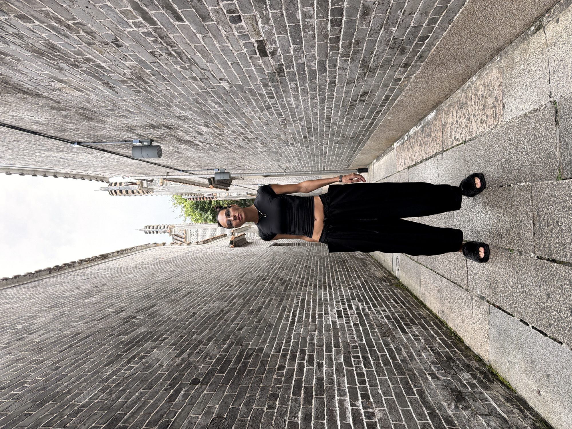 A full-body, medium shot shows a woman standing in a narrow alleyway in Chancheng District, China. The alley is paved with grey stone slabs and flanked by tall, dark grey brick walls. The woman, wearing a black cropped t-shirt, loose black pants, and black fluffy slides, stands facing the camera. She has dark hair pulled back and is wearing sunglasses. She is standing on the left side of the alleyway. The alley extends into the distance, with glimpses of traditional Chinese architecture with tiled roofs visible at the far end. A grey electrical conduit runs along the right wall. The sky is overcast.