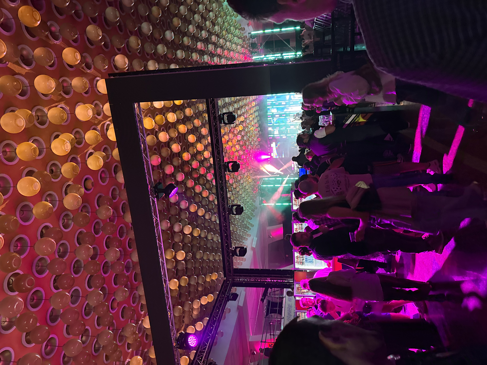 The image shows a concert venue in Yerevan, Armenia. A large crowd of people are gathered to watch a performance on stage. The stage is lit with bright pink and green lights. The walls of the venue are covered with rows of round lights. A black metal frame hangs above the stage with additional lights attached to it. The audience members are standing and watching the stage. Some people are holding drinks. The lighting in the venue is predominantly pink and purple.