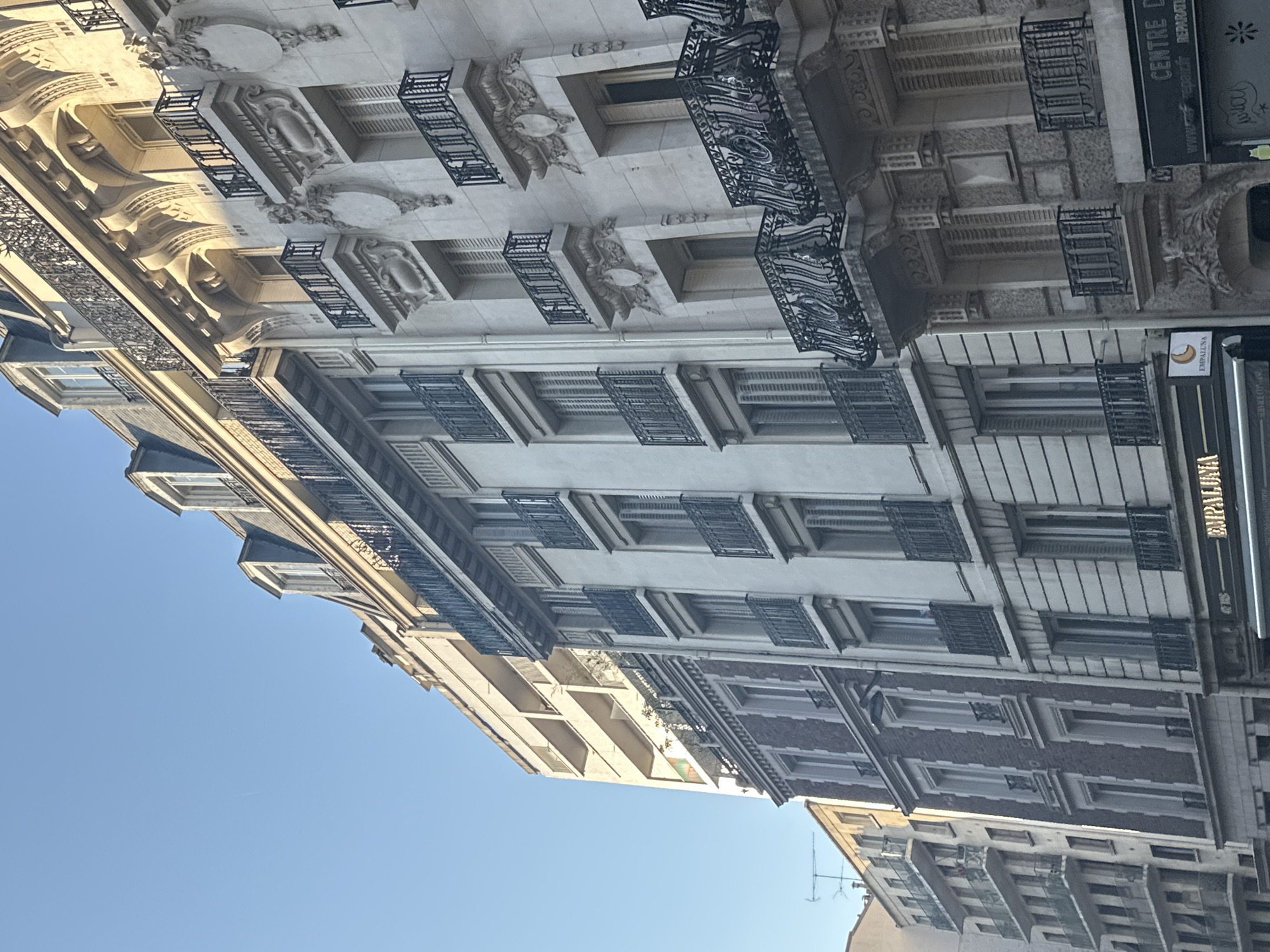 The image captures a view of Parisian architecture under a clear blue sky. The buildings are tall and multi-story, with distinct architectural styles visible. On the right, a building displays ornate stonework, decorative balconies with wrought-iron railings, and shuttered windows. Adjacent to it, a building with a brick facade features tall, narrow windows and balconies. Further to the left, a more modern building with a light-colored facade shows multiple levels of balconies. At street level, signage for "CENTRE DE" and "EMPALUNA" is visible. The scene suggests an urban environment in Paris, France, likely a residential or mixed-use area, with a focus on the building facades and their architectural details.