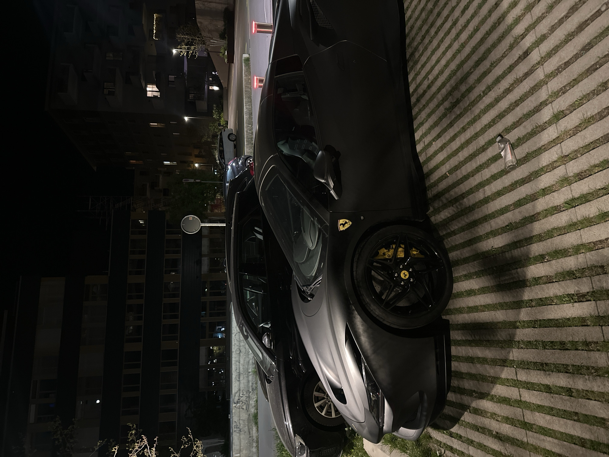 The image displays a dark-colored Ferrari sports car, likely matte black or dark grey, parked at night in an urban or suburban setting in Dübendorf, Switzerland. The car is positioned on a paved surface composed of striped concrete pavers with grass growing in the gaps. Its black multi-spoke wheels showcase bright yellow brake calipers and a yellow Ferrari emblem on the wheel hub, matching a larger emblem on the front fender.
In the immediate vicinity of the Ferrari, a clear plastic bottle with unreadable white and red text lies on the ground. Another lighter-colored car is partially visible behind the Ferrari. The background is dominated by several dark, multi-story buildings, some with illuminated windows, suggesting residential or office blocks. Streetlights and red-glowing bollards provide additional artificial illumination. A small, round mirror on a pole is visible on the far left. The sky is dark, indicating nighttime, and no people or specific activities are taking place; the cars are stationary. The scene appears dry and still.