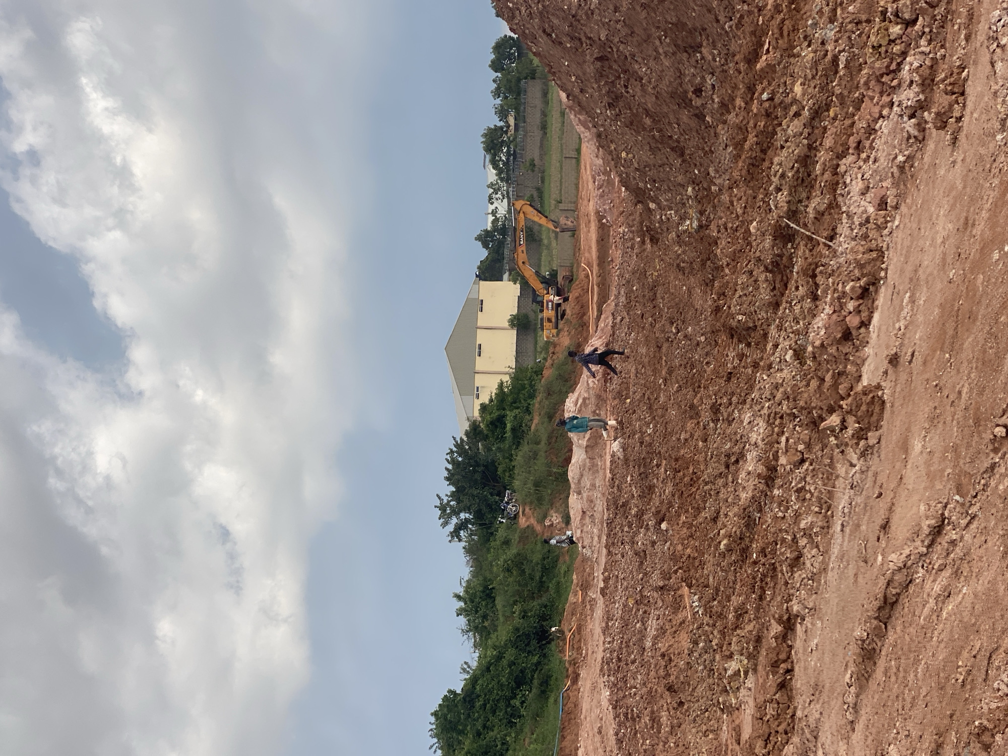 This image captures an outdoor construction scene in Reifild-Ist, Nigeria. The foreground is dominated by a steep, reddish-brown dirt embankment, indicative of ongoing excavation or earthworks. A few people are visible on and around the embankment, engaged in what appears to be construction labor. One person is standing on the higher part of the embankment, while others are lower down, near the base of the cut.
In the middle ground, an excavator, identified by the text "SANY" and "SY135C", is positioned in a trench, actively digging or moving earth. Beside the excavator, a yellow hose snakes across the ground. Further back, a light-colored, rectangular building with a visible roofline stands amidst lush green trees. The sky is partly cloudy with diffused sunlight, suggesting it is daytime, possibly late morning or afternoon, under a generally fair but overcast sky. The overall impression is one of a busy, ongoing construction project in a somewhat rural or semi-developed area.