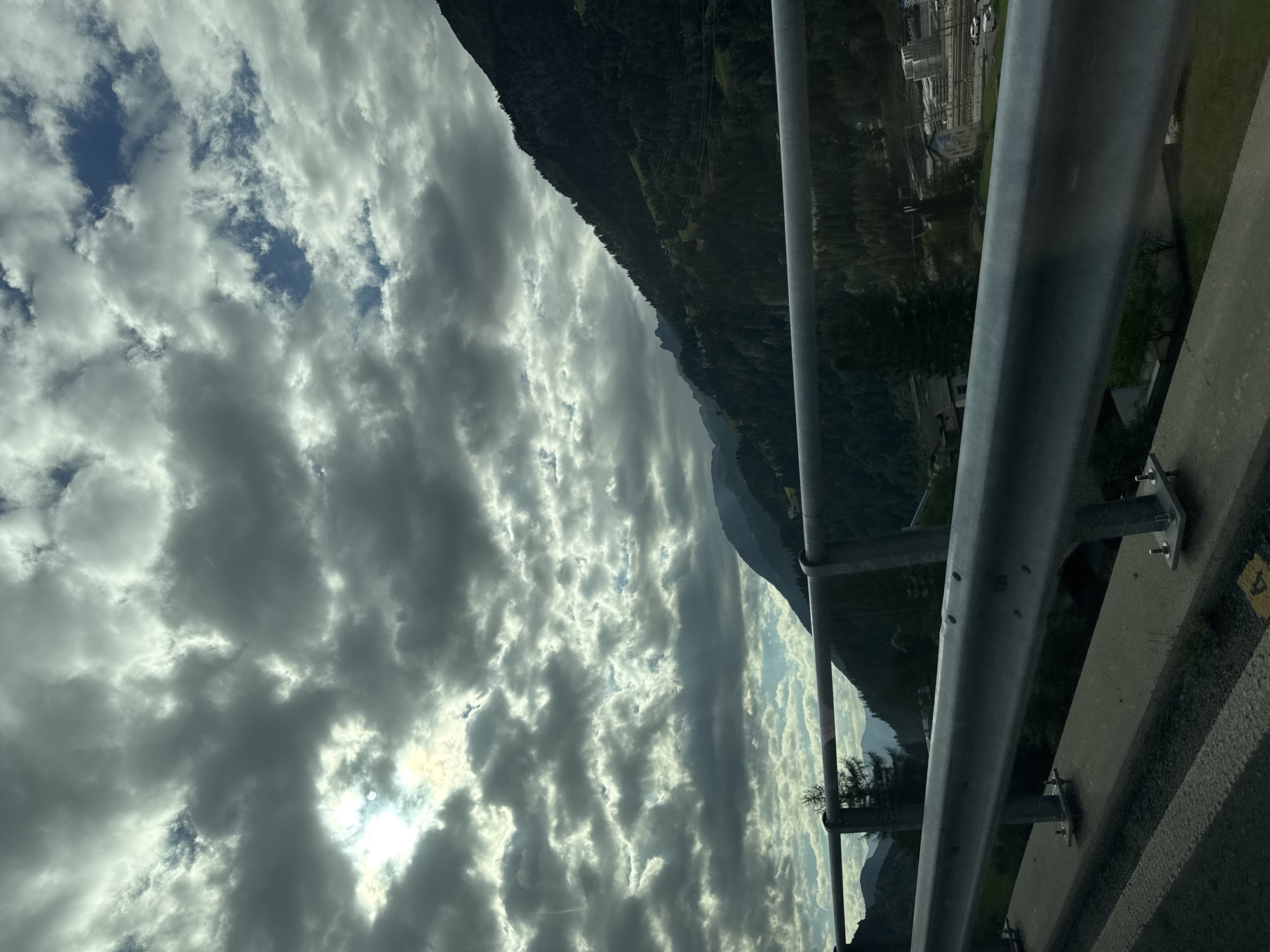 The image captures a mountainous landscape with a cloudy sky, viewed from a roadside barrier. The foreground shows a silver metal guardrail with visible bolts, mounted on concrete supports. Beyond the guardrail, a densely forested mountain slope dominates the right side of the frame, with a winding road visible in the distance, featuring scattered buildings and some vehicles. The sky is filled with dramatic, heavy clouds, with patches of sunlight breaking through, creating a dynamic lighting effect. The scene appears to be in the vicinity of Airolo, Switzerland, a town known for its mountainous terrain and picturesque views. There are no people or specific actions depicted in the image. The overall environment is natural and rugged, characteristic of an alpine region.