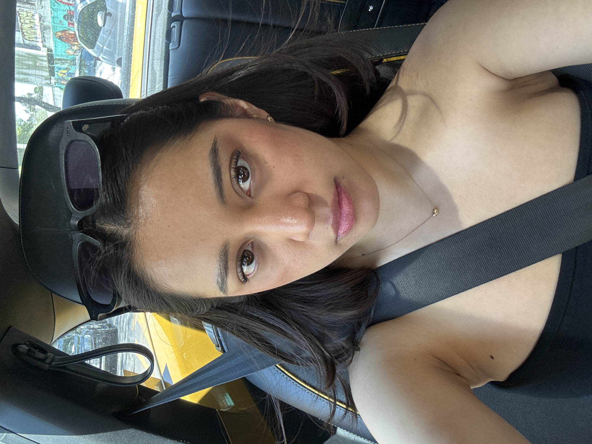 A mid-angle, close-up indoor shot shows a woman with long, dark hair and brown eyes, wearing a seatbelt in what appears to be a taxi cab. Sunglasses are on her head. She is wearing a gold necklace and a dark, possibly black, top. A portion of the window is visible, suggesting an urban landscape of Mexico City, Mexico. Graffiti and building structures are discernible through the window. The car's interior has black leather seats and accents of yellow.