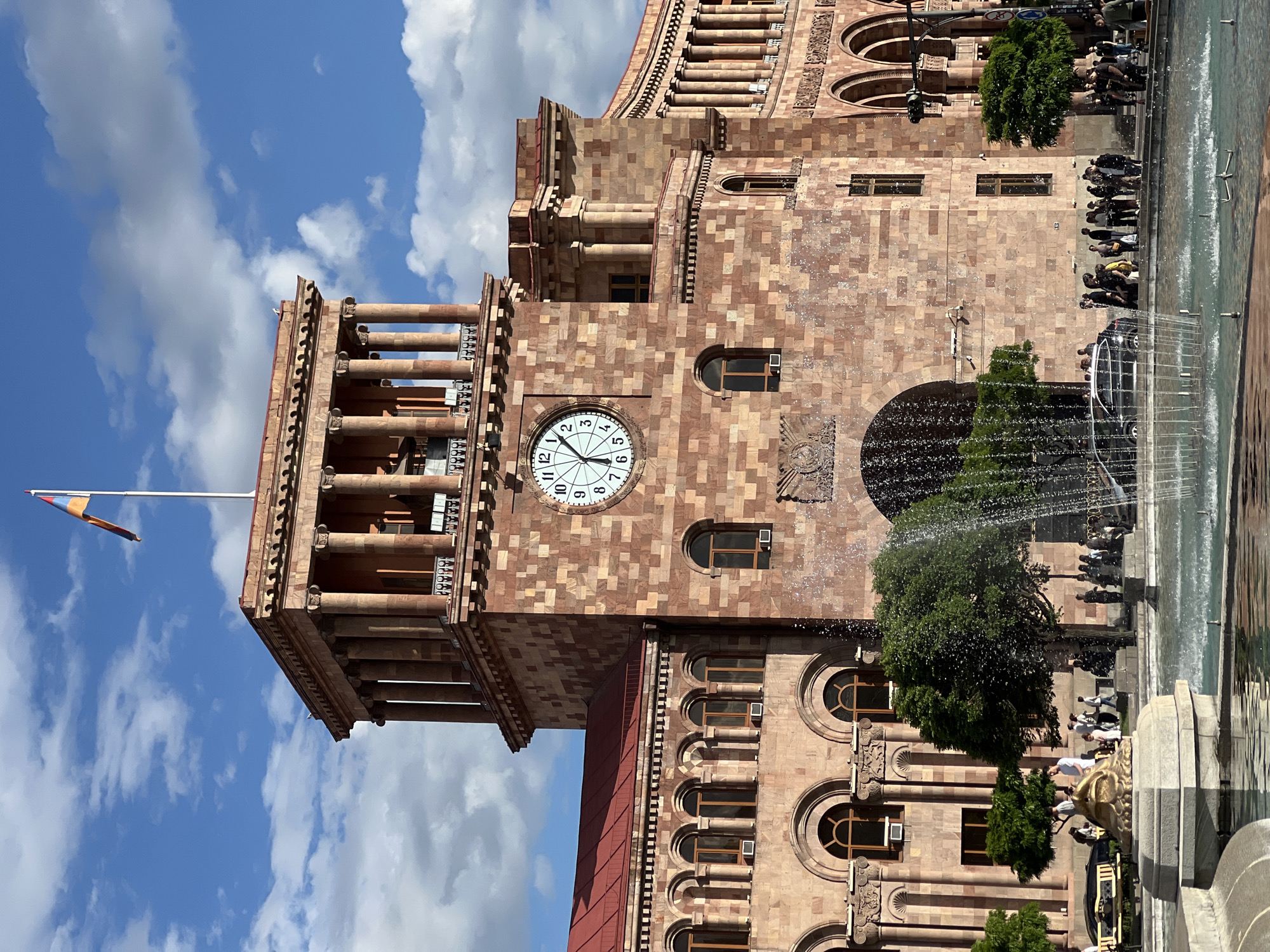 The image is an upward-angled shot of a red stone building with a clock tower, located in Yerevan, Armenia. The clock on the tower reads approximately 7:06. The building's facade features rows of windows and decorative columns. A flag is attached to the top of the clock tower.
In the foreground, a fountain with water jets spraying is partially visible. Pedestrians are present near the fountain. The sky above is blue with white clouds.
