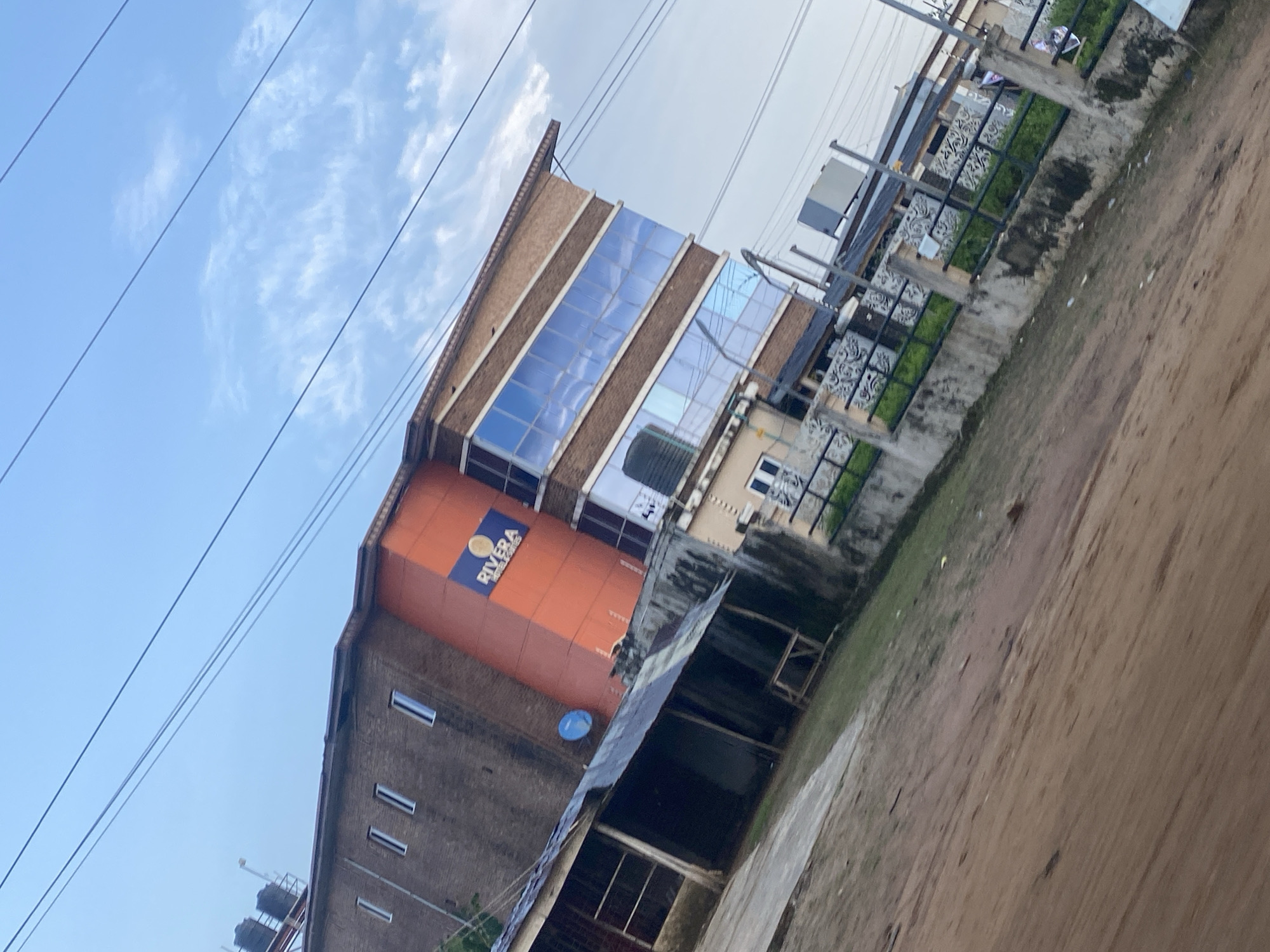 The image shows a building with the sign "RIVERA HOTEL & SUITES" located in Ikorodu, Nigeria. The building is multi-story with a brick facade and large blue-tinted windows. Part of the building is painted orange. In front of the building, there is a smaller, older structure with a corrugated metal roof. A dirt road is in the foreground, and a concrete fence with decorative metalwork borders the property. Overhead, power lines are visible against a cloudy sky. The scene appears to be an outdoor view of a commercial establishment.
