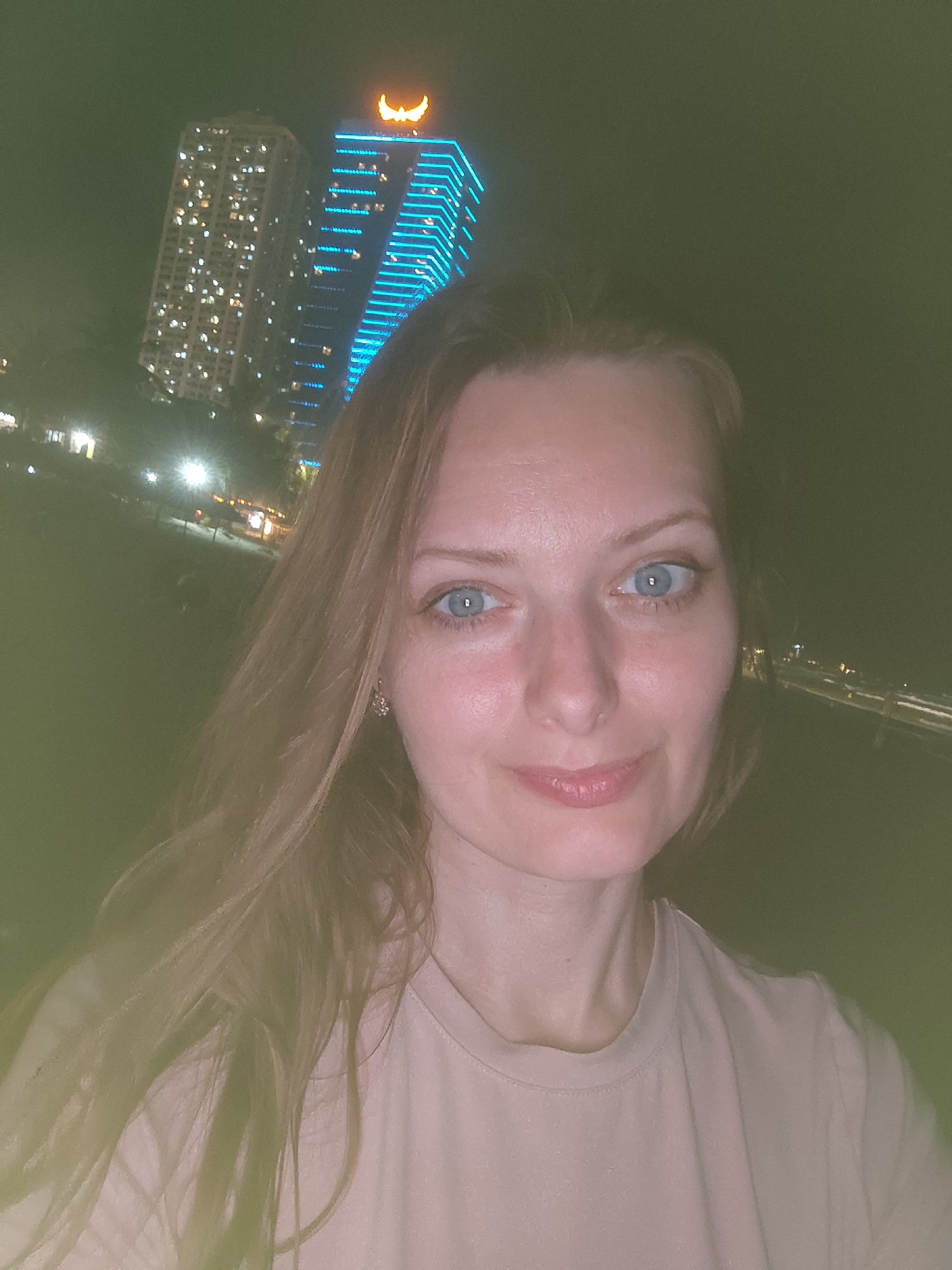 A medium shot of a woman at night. She is fair-skinned with blue eyes and long, light brown hair. She is wearing a light pink t-shirt. Her expression is relaxed and she is looking directly at the camera. In the background are two skyscrapers, one illuminated with blue neon lights. The skyline and streetlights behind the skyscrapers suggest that the picture was taken in a city. Based on the building with the blue neon lights, this image was most likely taken at night along the Han River in Đà Nẵng, Vietnam. The building in the background is the Novotel Danang Premier Han River hotel.