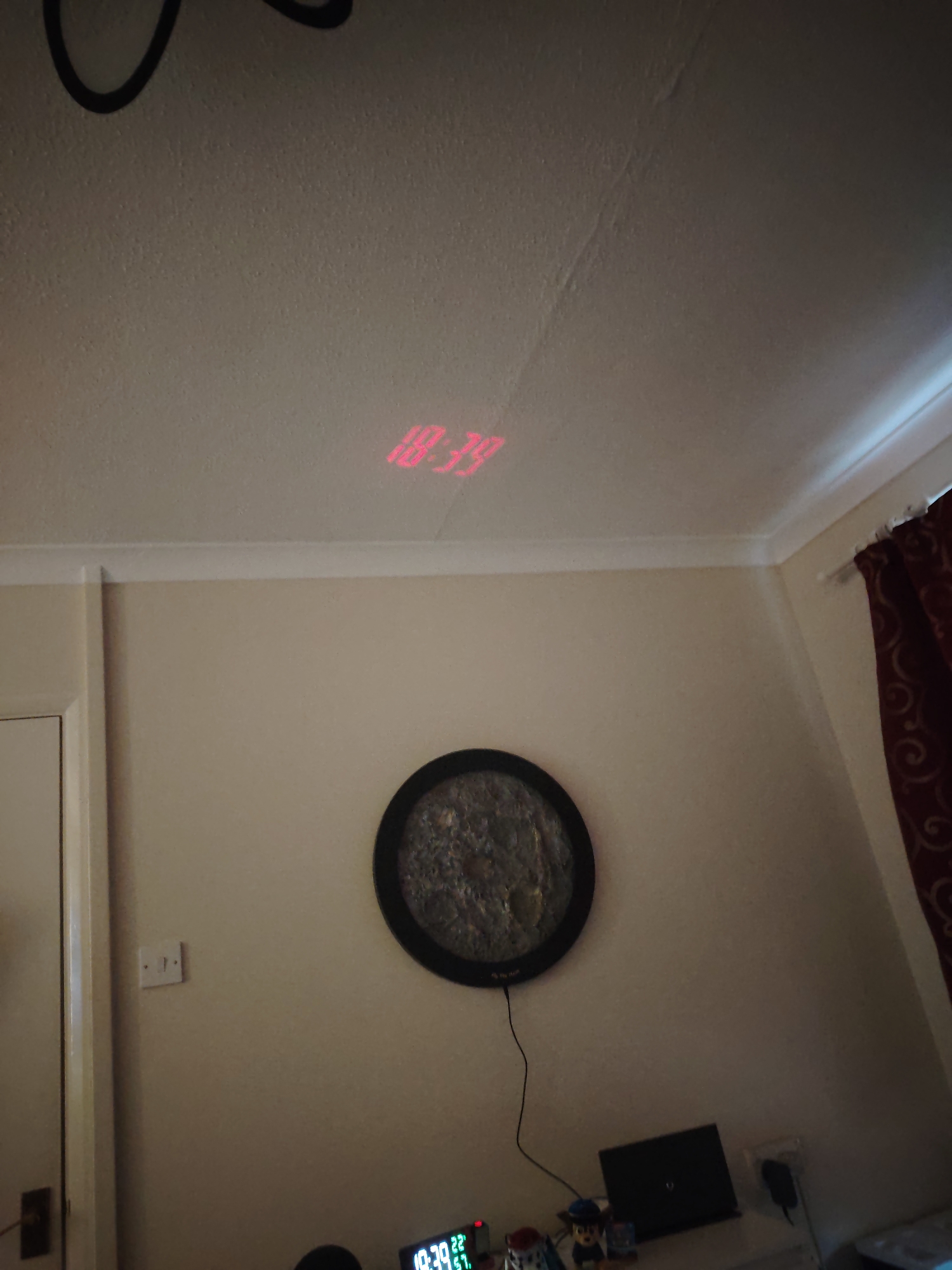 This image captures an indoor scene, likely a bedroom or study, in Huntingdonshire, United Kingdom. The room features light-colored walls and a textured white ceiling, displaying visible cracks or imperfections.
The most prominent detail is a red digital time, "10:39", projected onto the ceiling. Below, a light switch is mounted on the wall to the left. Centered on the wall is a dark-framed, circular piece of art or decoration, resembling a textured lunar surface, with a black power cord extending downwards. Red patterned curtains are visible on the right side of the frame.
On a surface at the bottom, a small digital alarm clock prominently displays "10:39", matching the projected time. Smaller green numbers on the clock show "22:51" and "20:41". Other objects on the surface include a closed black laptop, a small speaker or electronic device, a white mug, a Paw Patrol figurine (specifically Chase, with "PAW" visible on his uniform), and various cables and a power adapter. The overall lighting is dim, indicating either early morning or, more likely given the use of a projection clock, evening or nighttime. No people are visible, and there is no direct activity taking place beyond the display of time.