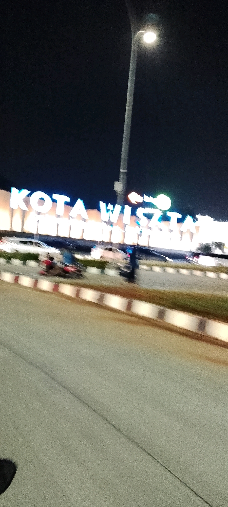 The image shows a blurry, night-time scene with a large, illuminated sign that reads "KOTA WISATA." The sign is composed of bright, white block letters with a blue outline. To the right of the sign, there is a green arrow pointing left, with a light bulb symbol. A tall, slender street lamp with a bright light at its top is visible on the right side of the image, illuminating the scene. In the foreground, a paved road is visible, with a grassy median strip separating it from the area where the sign is located. Cars and a motorcycle are blurred in motion on the road and the median. The overall environment suggests a well-lit area, likely an entrance or prominent landmark within the "Kota Wisata" development.