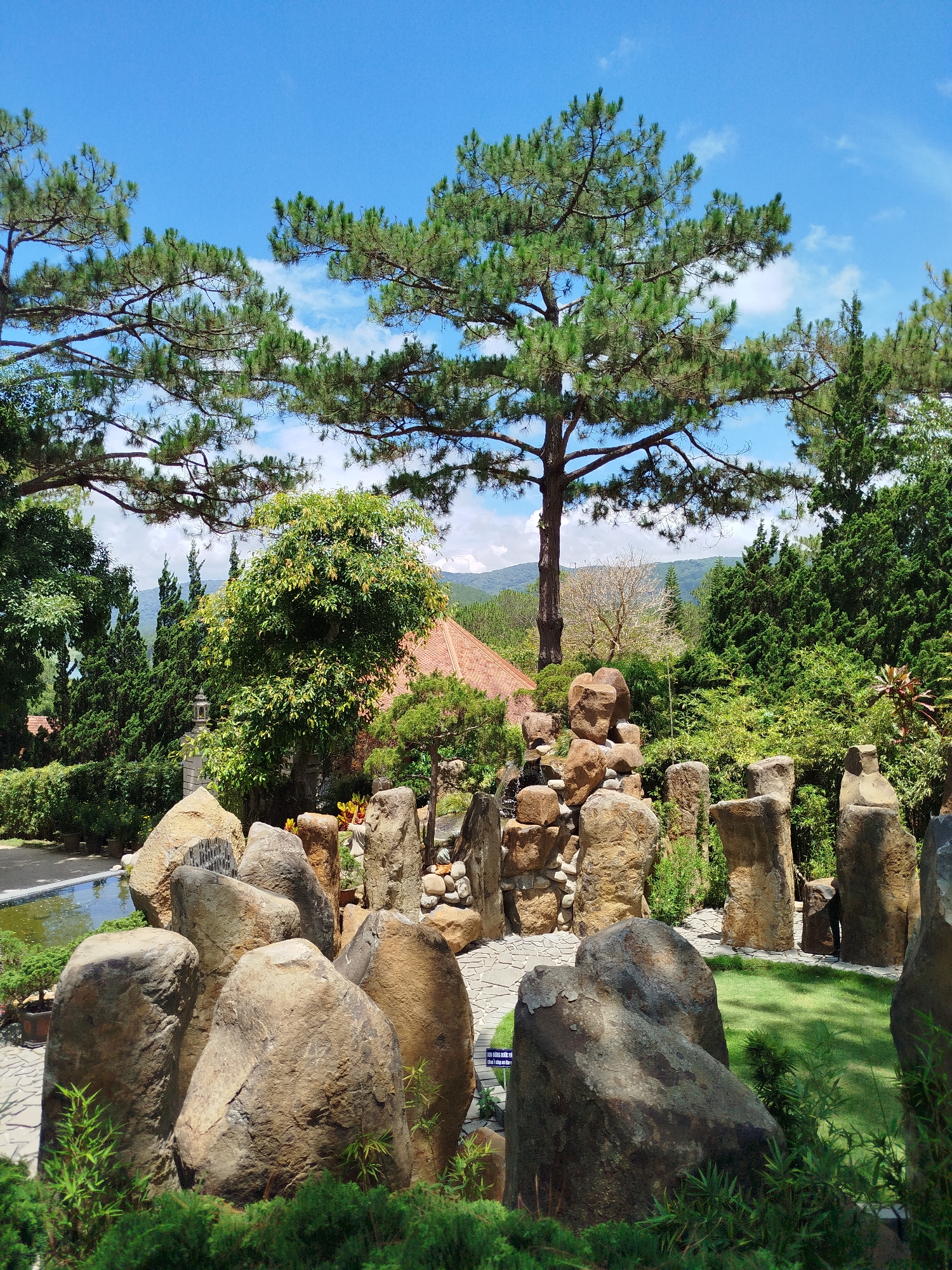 The image shows a landscaped garden in Da Lat, Vietnam. Dominating the foreground are large, irregularly shaped brown rocks and smaller stones interspersed with green shrubbery. A narrow stone-paved path leads between these rocks. In the mid-ground, a small waterfall cascades down a rock structure. Behind this, the garden transitions into lush greenery including various trees and plants. A red-tiled roof of a building is partially visible among the foliage. A tall pine tree stands prominently. In the background, distant hills are visible under a bright blue sky with scattered white clouds. No people are visible.