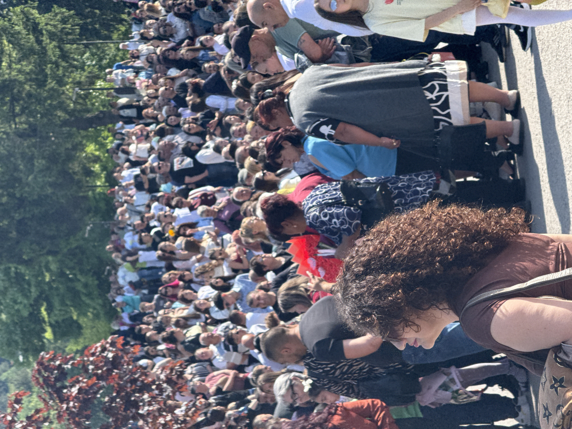 A dense crowd of people occupies a space beside a road, likely gathered to watch or participate in an event. The scene is in the city of Shumen, Bulgaria. The crowd stretches into the distance, with many faces visible, most looking in the same direction. Varying attire is observed among individuals, including casual clothing. To the left, a line of trees with red leaves borders the crowd. The composition of the image suggests it was taken from within the crowd, closer to the front, with a clear view of a large segment of the gathering.
