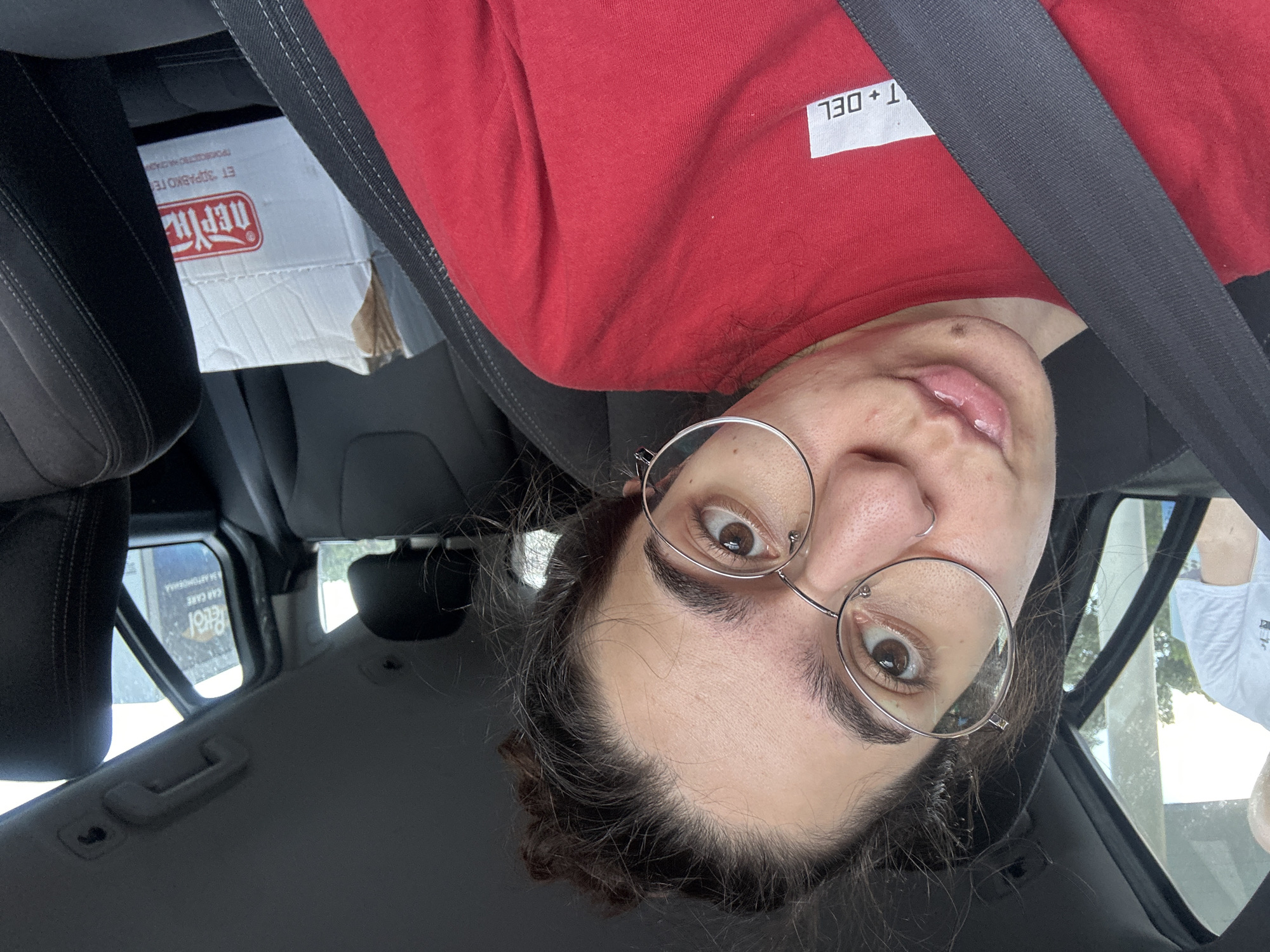 A young woman with dark curly hair pulled up in a bun and wearing round wire-rimmed glasses and a red t-shirt is sitting in the passenger seat of a car. She is looking at the camera with her mouth pursed and has a nose ring. A black seatbelt is visible across her chest. In the background, through the car window, a man in a white t-shirt is visible, as is a sign that reads "Petrol" and "CAR CARE". A cardboard box with Cyrillic lettering is on the seat next to her. The scene appears to be in a car parked at a petrol station or car wash in Sofia, Bulgaria.