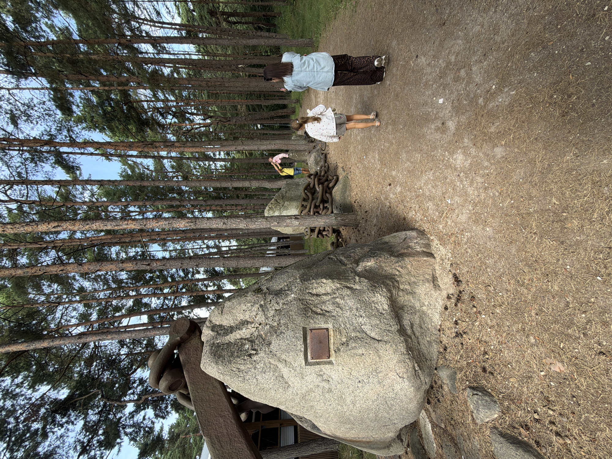 The image depicts an outdoor scene in a forested area, likely a park or trail, within the city of Ventspils, Latvia. In the foreground, a large granite boulder with a metal plaque is prominently displayed. To the right of the boulder, a dirt path meanders through the trees. Two individuals are walking away from the camera on this path: a young girl wearing a white patterned jacket and shorts, carrying a stuffed animal, and an adult woman in a light blue jacket and patterned pants. Further in the background, another group of people is visible interacting with large, rusty metal chains and another boulder. The environment is characterized by tall, slender pine trees and a ground covered in pine needles and cones. The overall atmosphere suggests a casual, recreational setting.