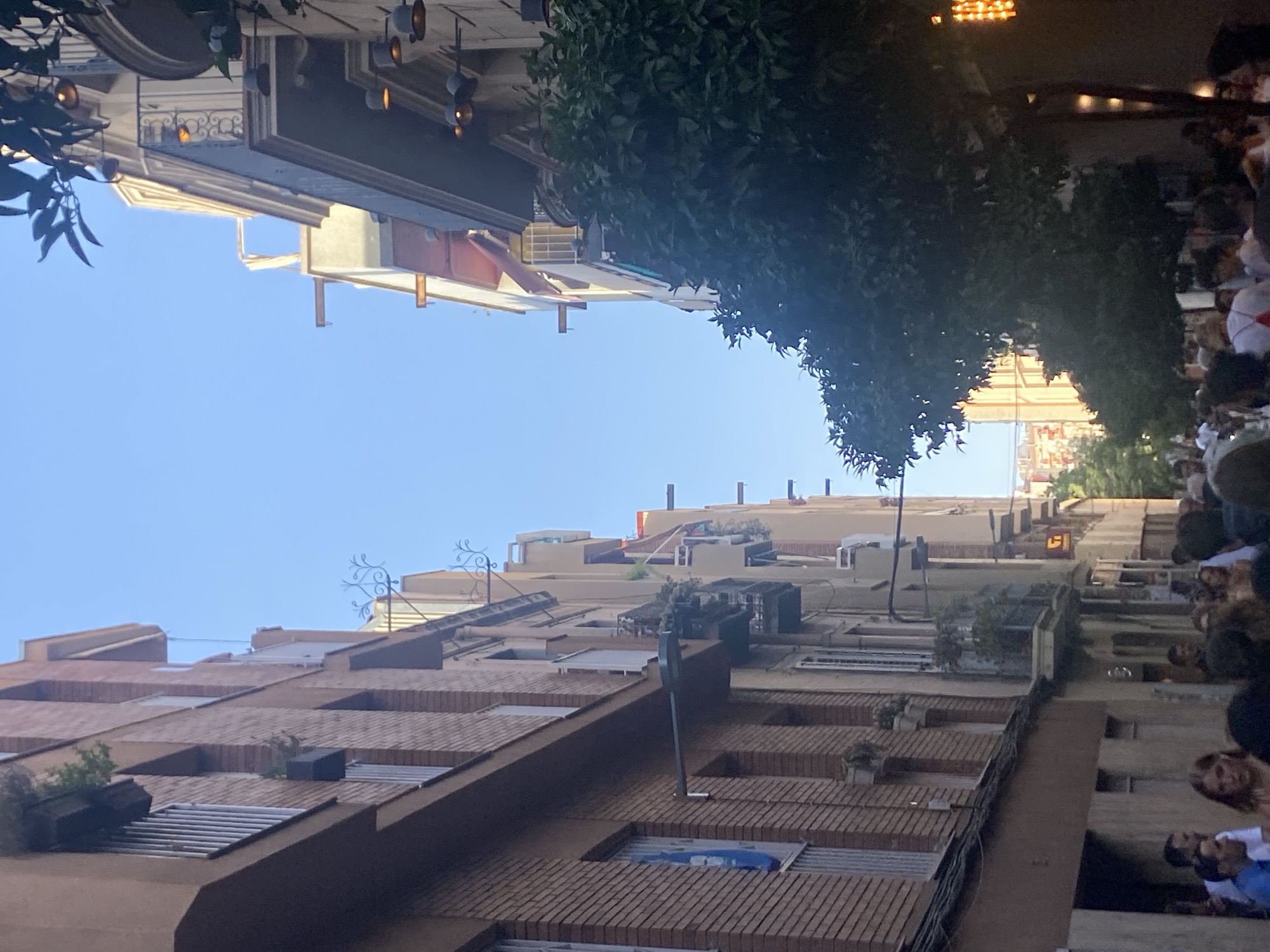 The image depicts a narrow street scene in Barcelona, Spain, with tall buildings lining both sides and a clear blue sky overhead. The architecture features brick facades on the left and a mix of light-colored plastered buildings on the right, with balconies adorned with plants and decorative railings. Outdoor lighting fixtures are visible on the building to the right, suggesting a commercial or residential area. A large tree with dense foliage partially obscures the view of the buildings on the right, adding a natural element to the urban landscape. A crowd of people is visible at the bottom of the image, walking along the street, indicating a busy atmosphere. Their attire suggests warm weather. The overall impression is of a vibrant street life in a European city.